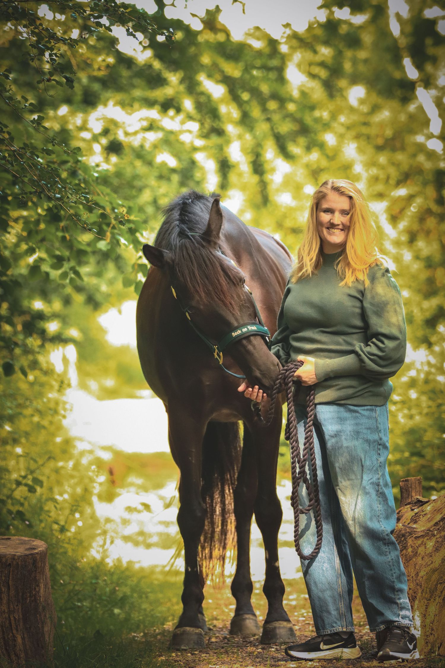 Smiling blonde woman standing with a dark brown horse on a leafy green tree-lined path, holding a rope lead.