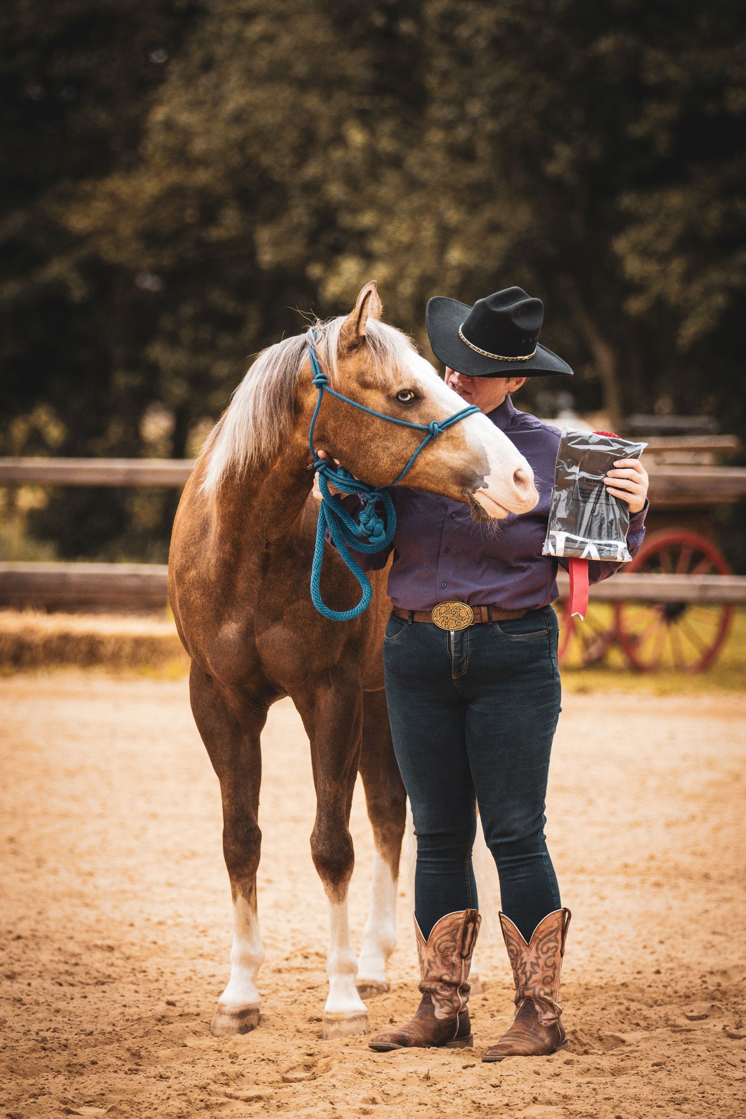 Cowgirl in black hat and boots embraces a chestnut horse while holding a trophy in a sandy arena.