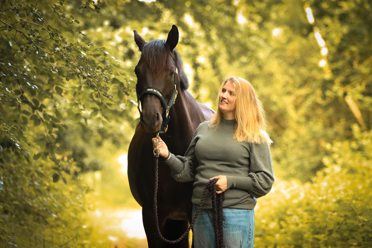 Woman with long blonde hair smiling beside a dark horse on a tree-lined path in lush green surroundings.