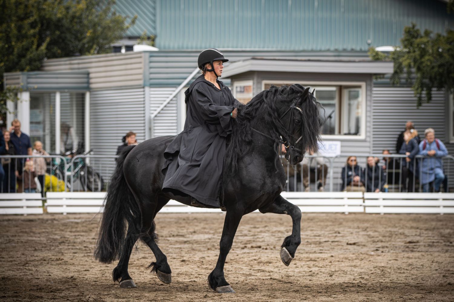 Rider in black cape performs dressage on a stunning black Friesian horse in an outdoor arena with spectators.
