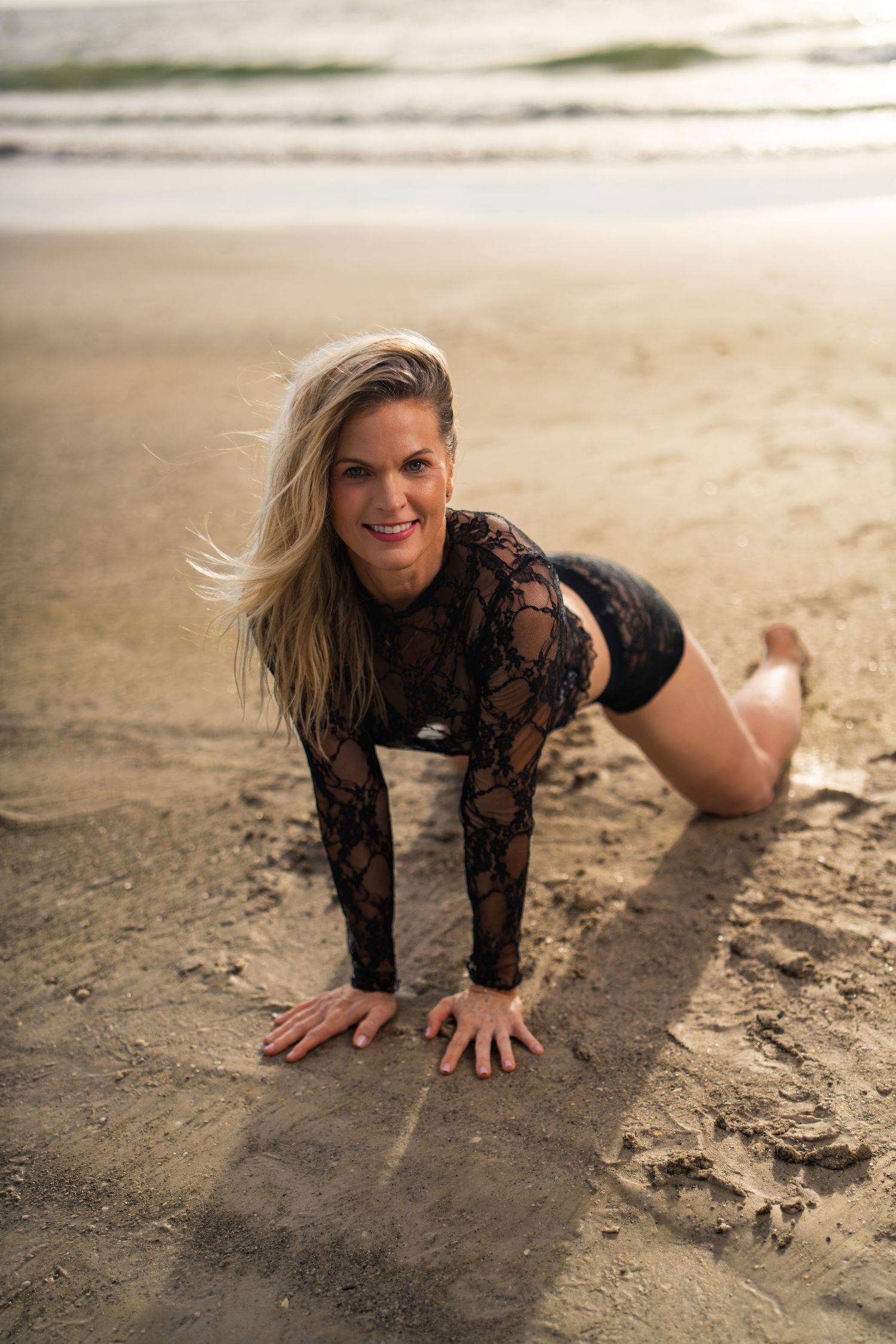 Woman in black lace outfit posing on sandy beach with ocean waves in background.