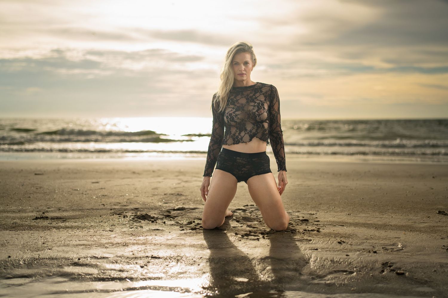 Woman in black lace top and shorts kneeling on wet beach sand at sunset with ocean waves behind her.