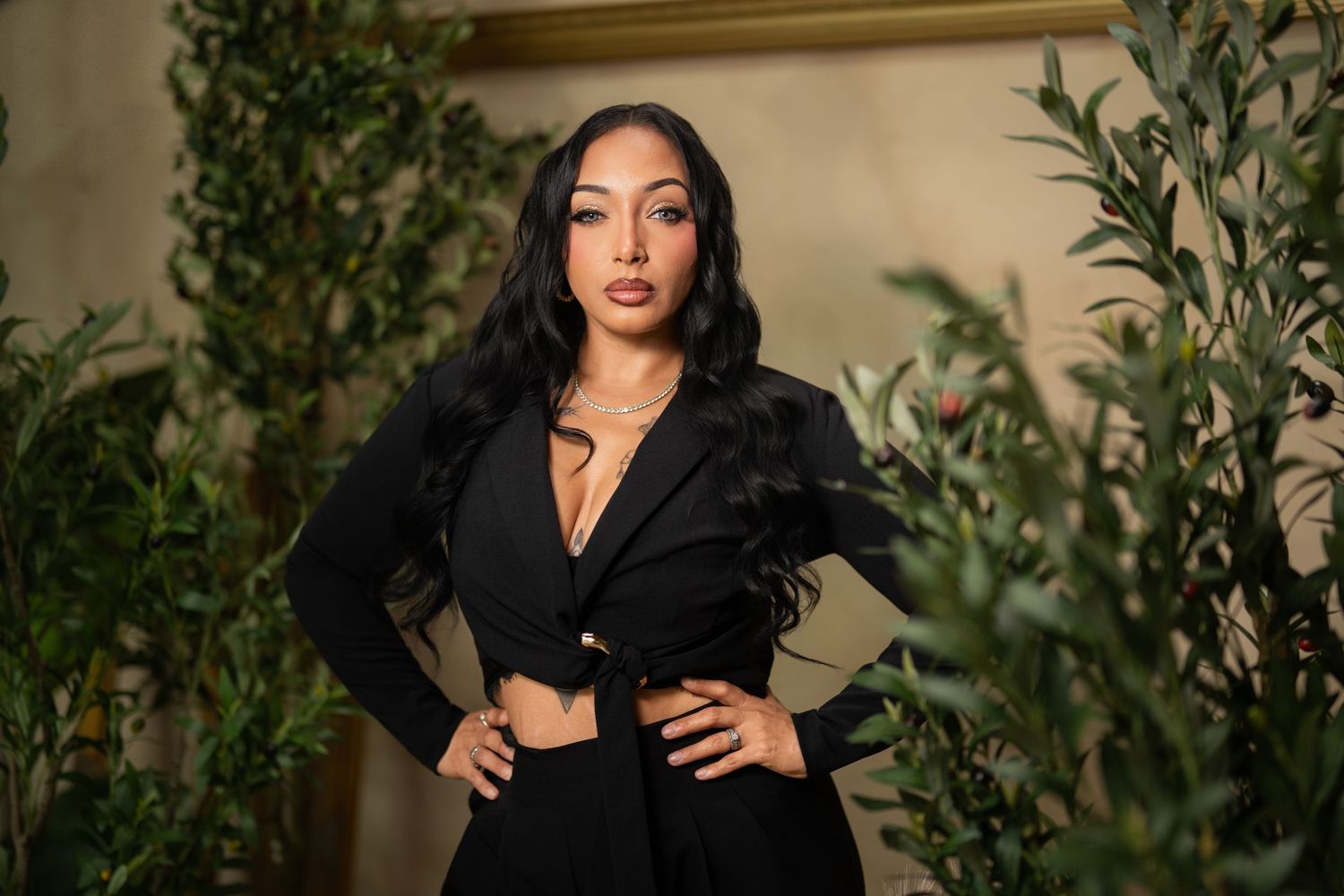 Woman with long dark wavy hair poses confidently with hands on hips in a black blazer and matching pants, framed by green olive branches.