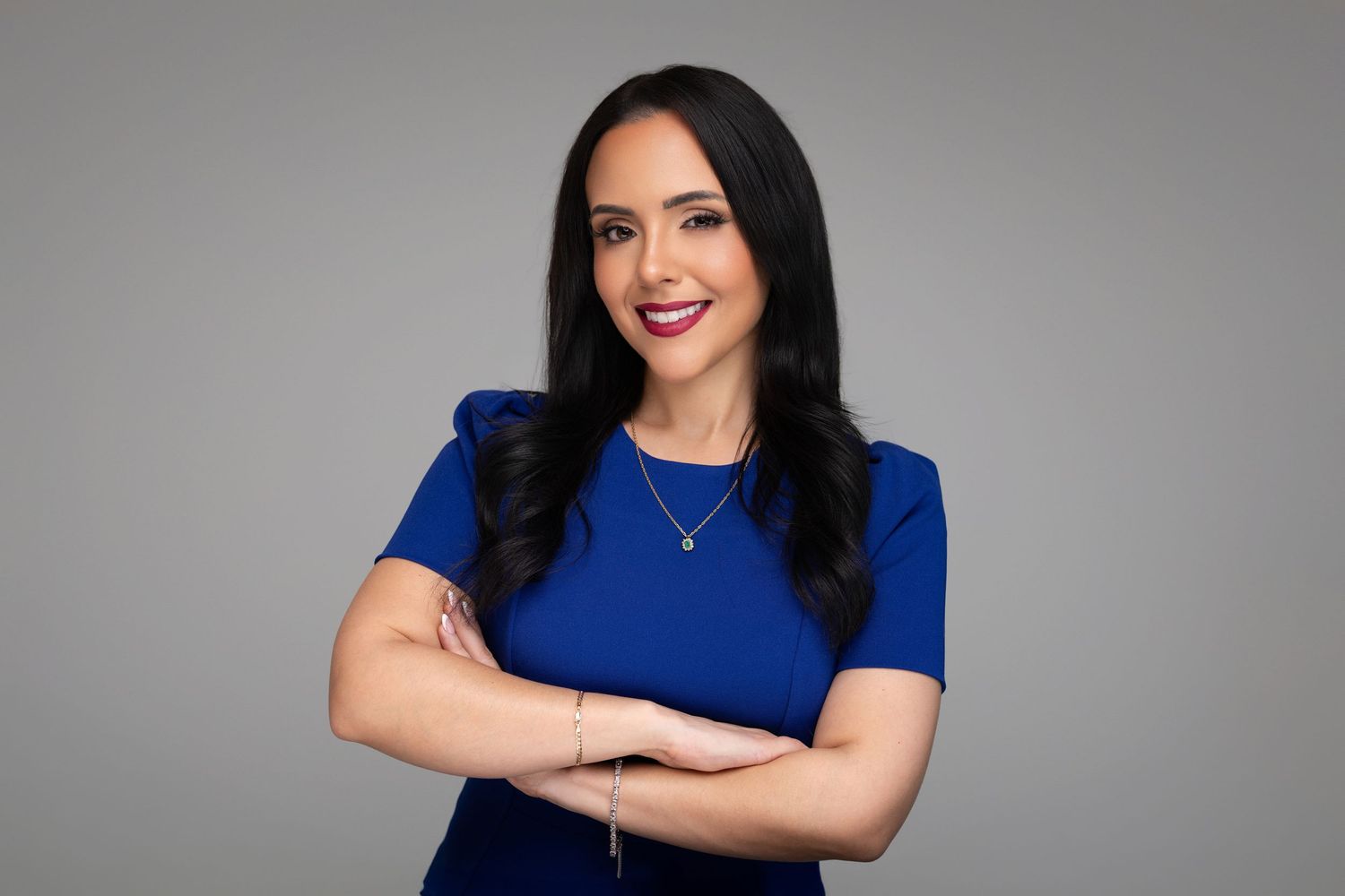 Smiling woman with long dark hair and red lipstick wears a royal blue dress and crosses her arms against a gray studio background.