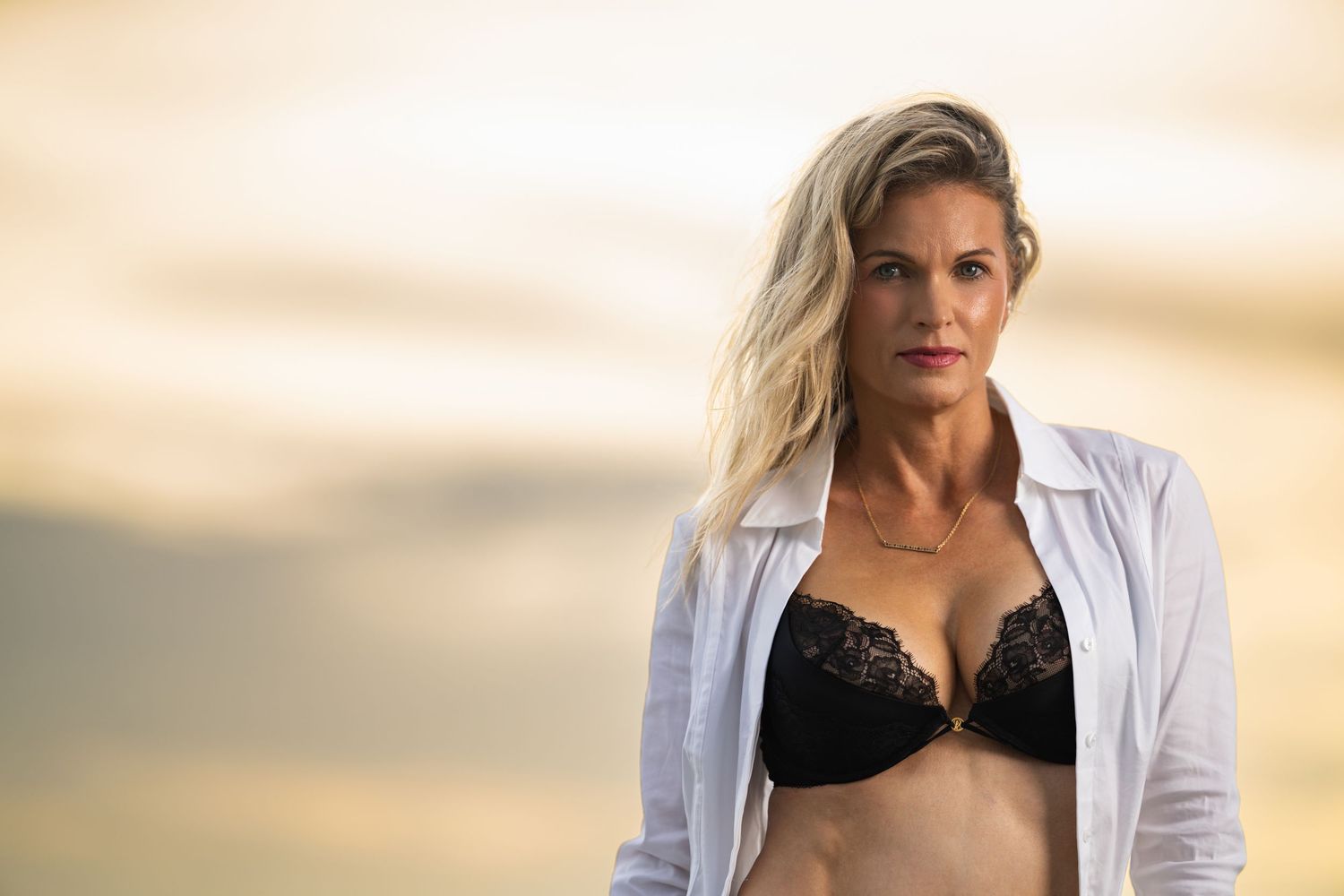 Blonde woman in black lace bra and open white shirt posing outdoors at sunset with golden sky background.