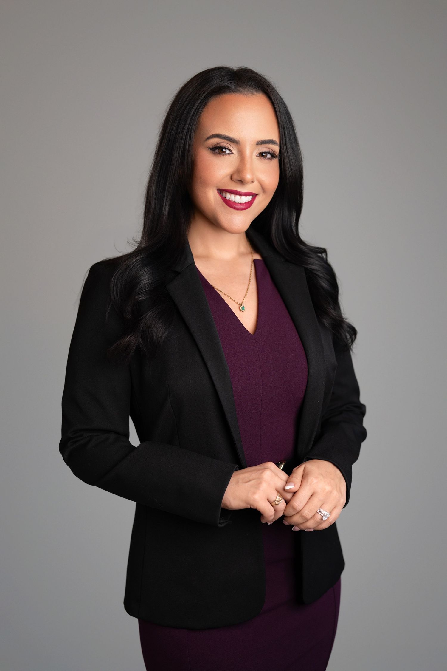 Smiling woman with long dark wavy hair wearing a black blazer over a plum dress, posing with hands clasped against a gray studio background.