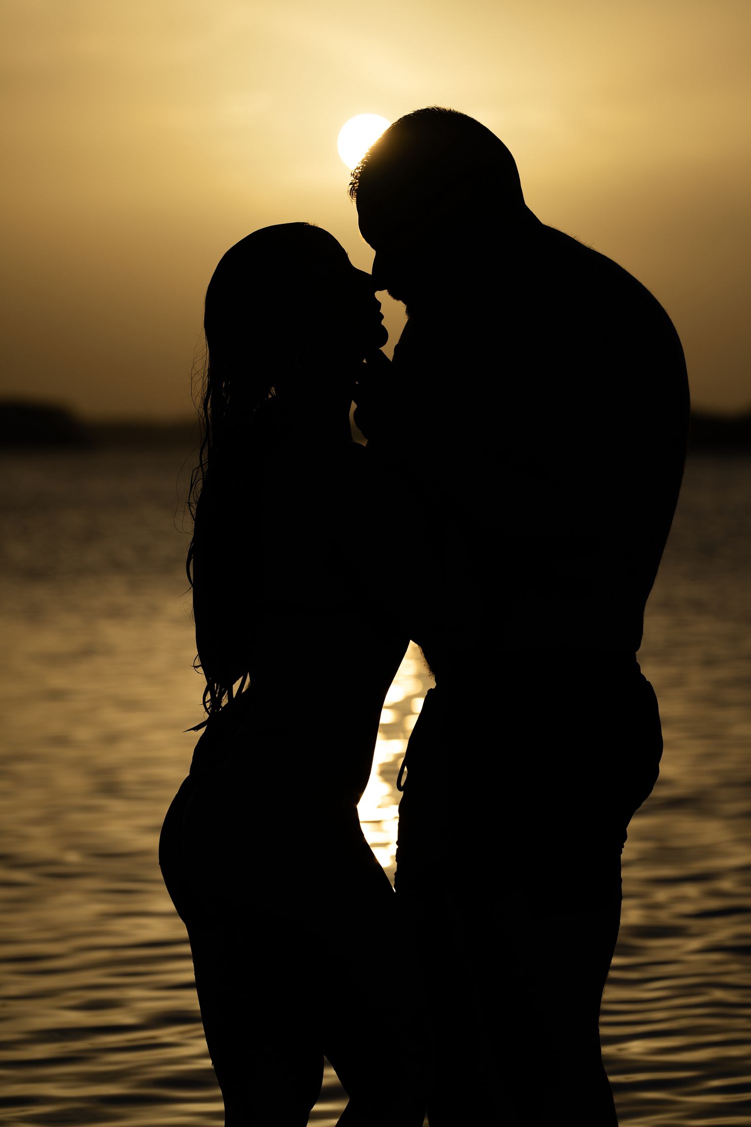 Romantic couple silhouette kissing at golden sunset by the water, backlit by warm glowing sun.