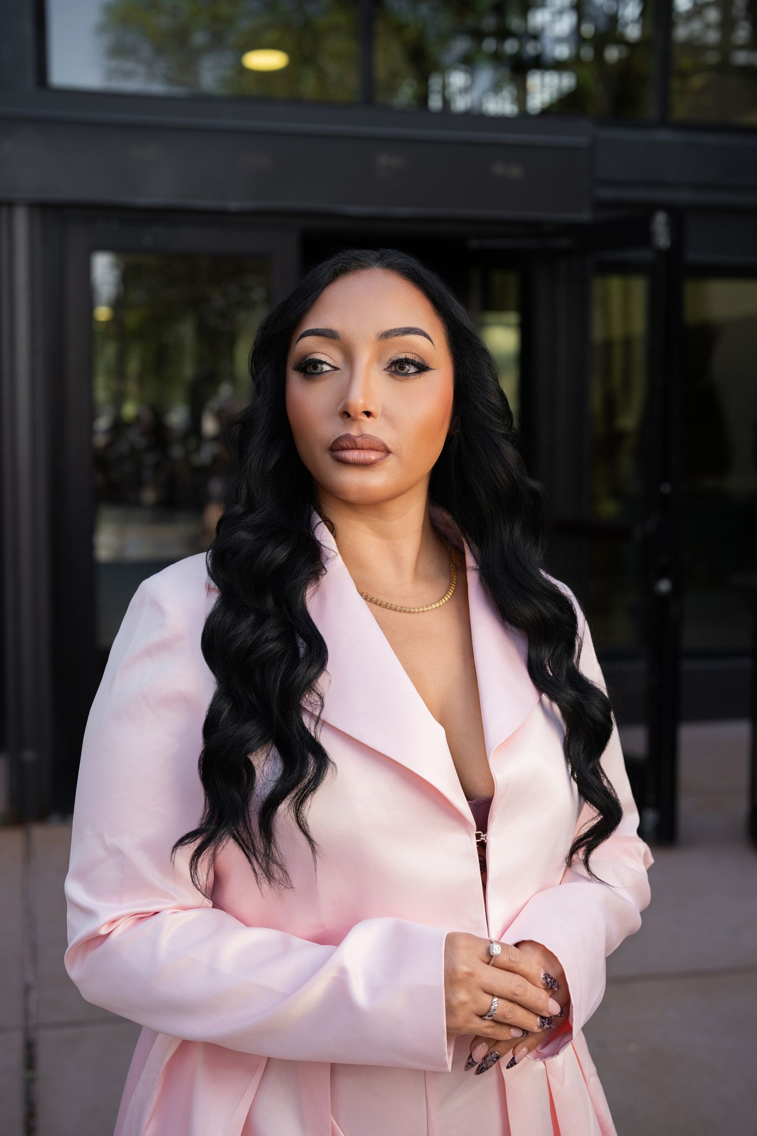 A woman in a pink blazer with long black wavy hair poses confidently outdoors near glass doors.