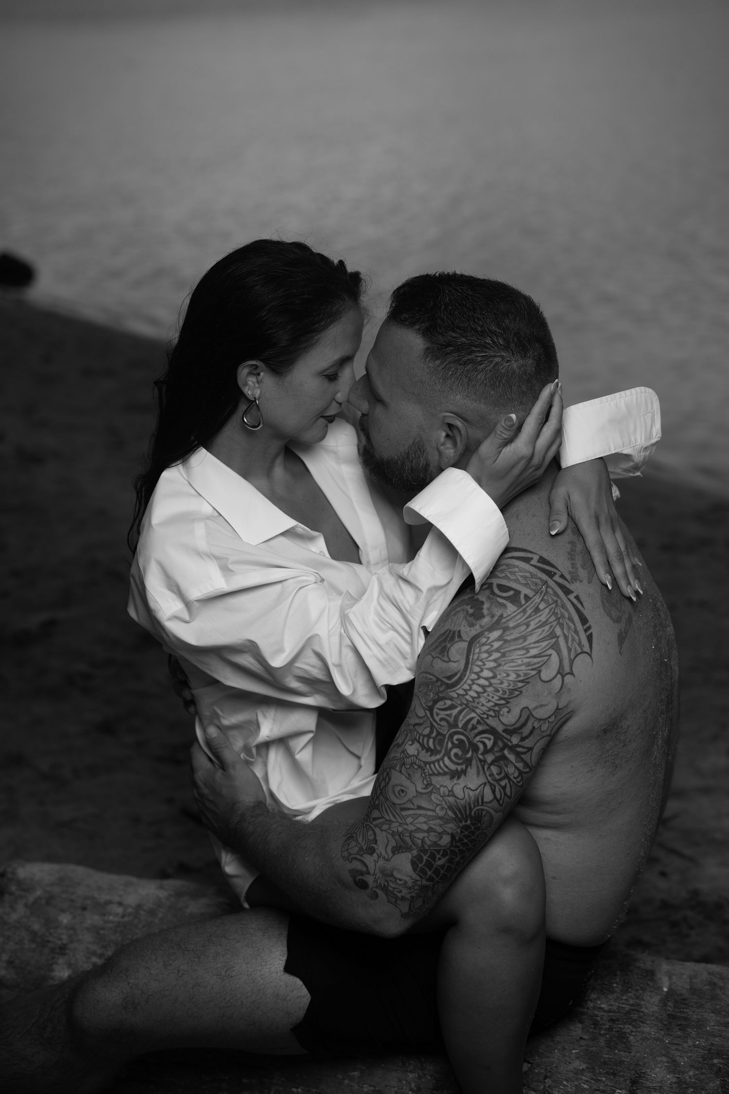 Black and white photo of a tattooed shirtless man embracing a woman in a white shirt near water.