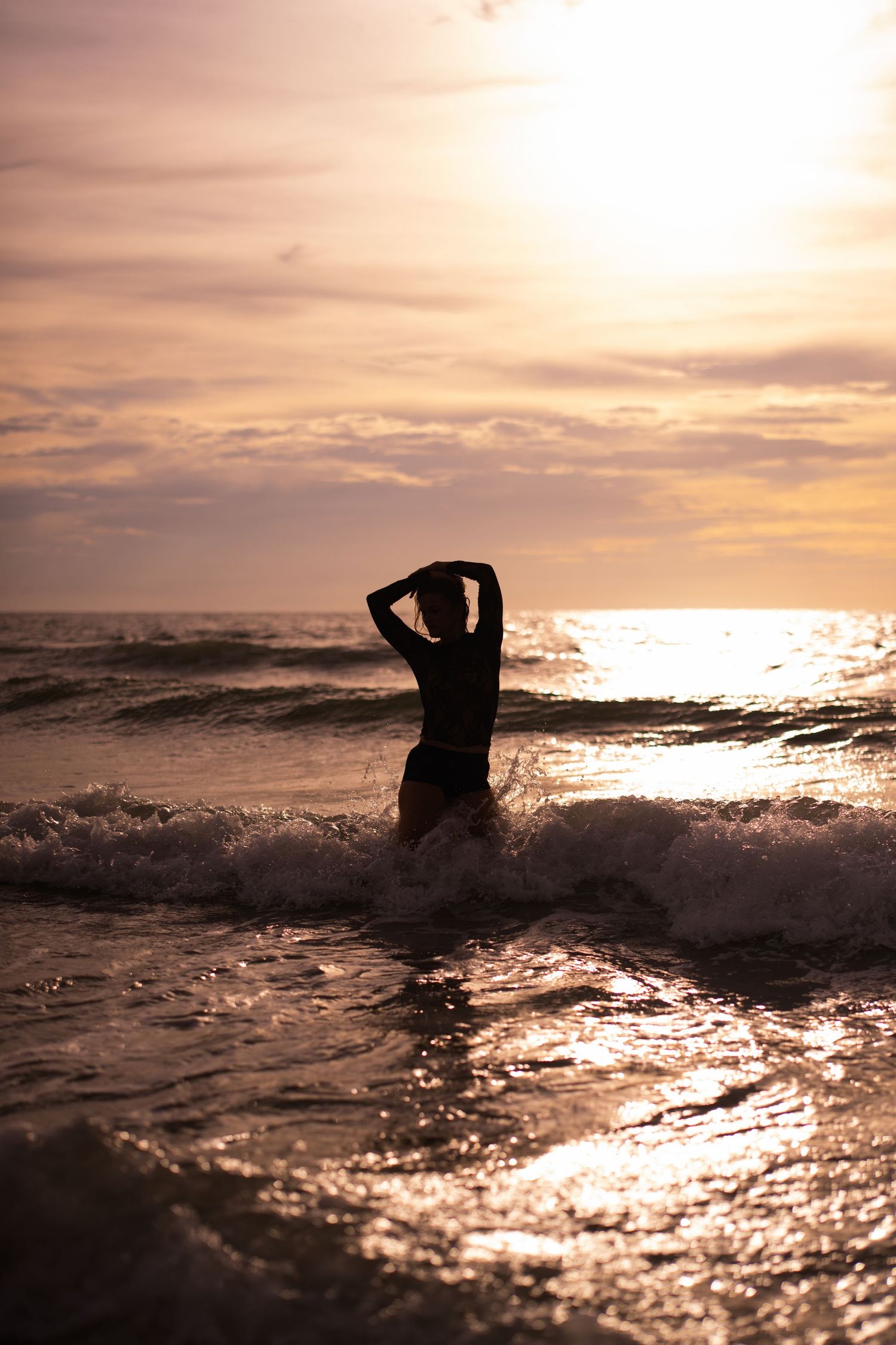 Silhouette of a woman standing in ocean waves during a golden sunset, with hands raised above her head.