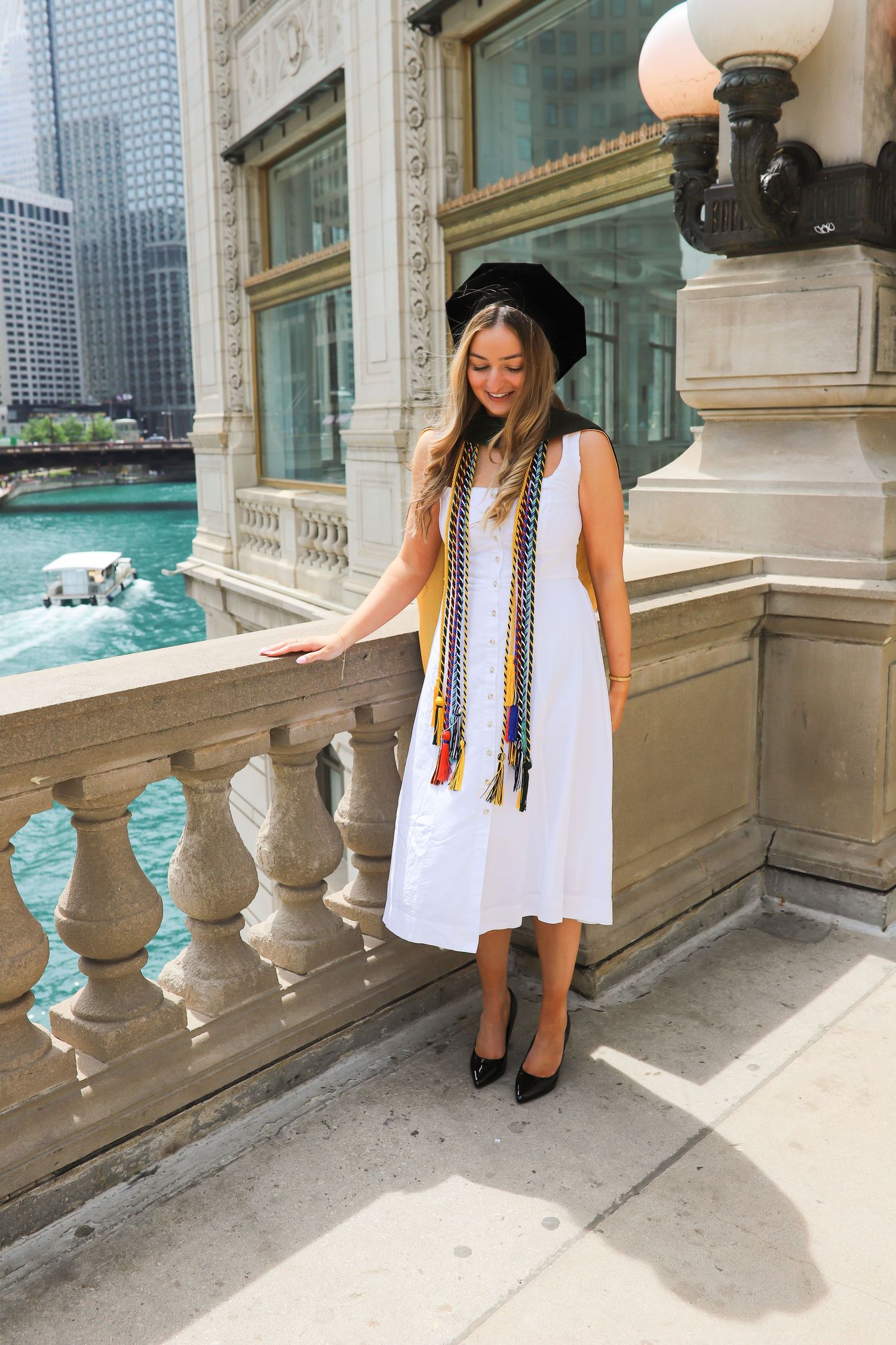 Young woman in white dress and graduation cap poses on ornate balcony overlooking a turquoise river in Chicago.