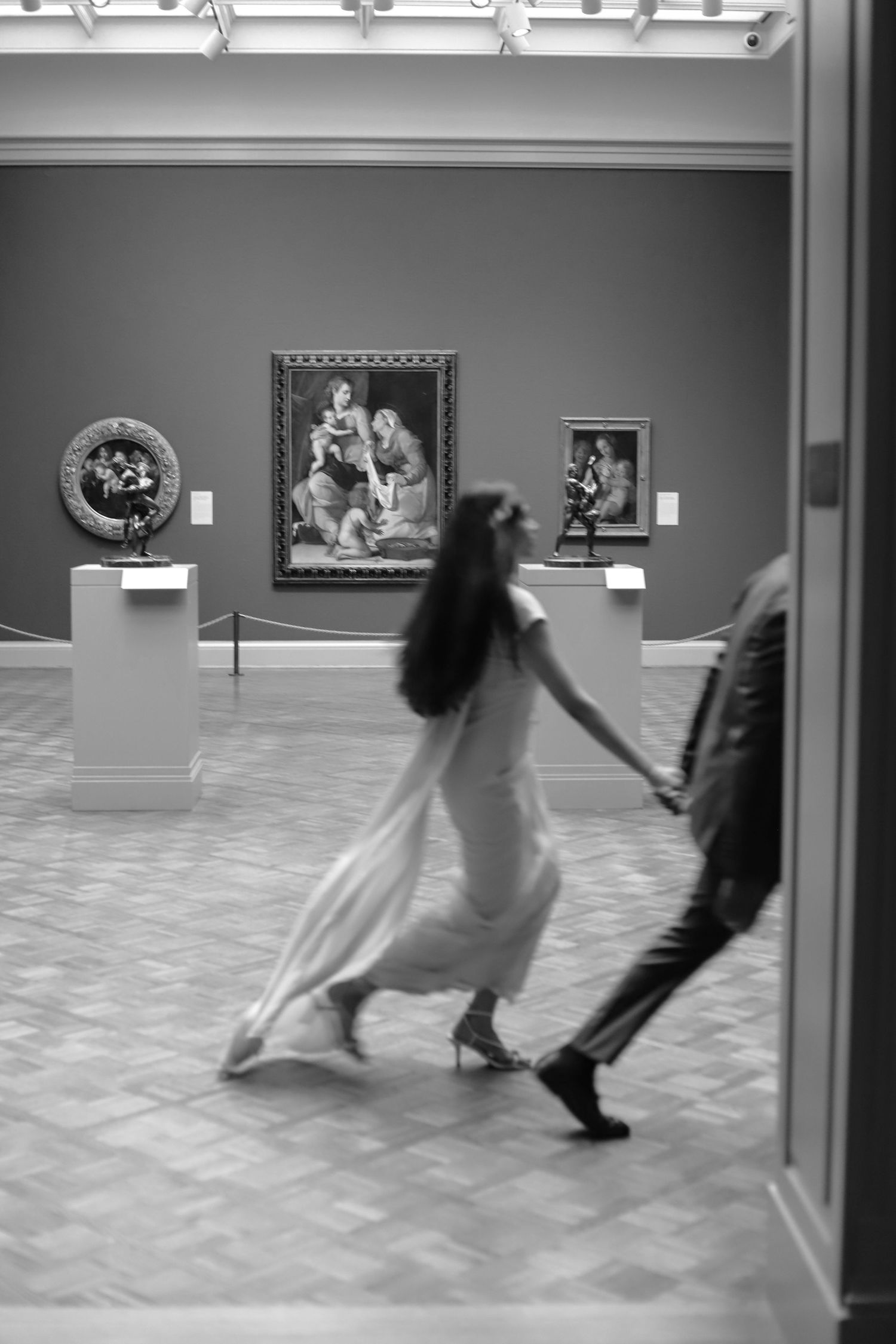 Black and white photo of a couple holding hands, walking through a classic art museum gallery.