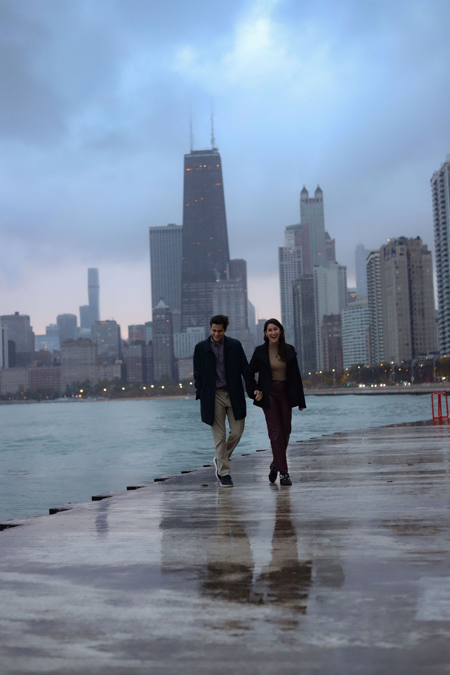 Couple holding hands walking along a wet lakefront pier at dusk with the Chicago skyline and moody overcast sky in the background.