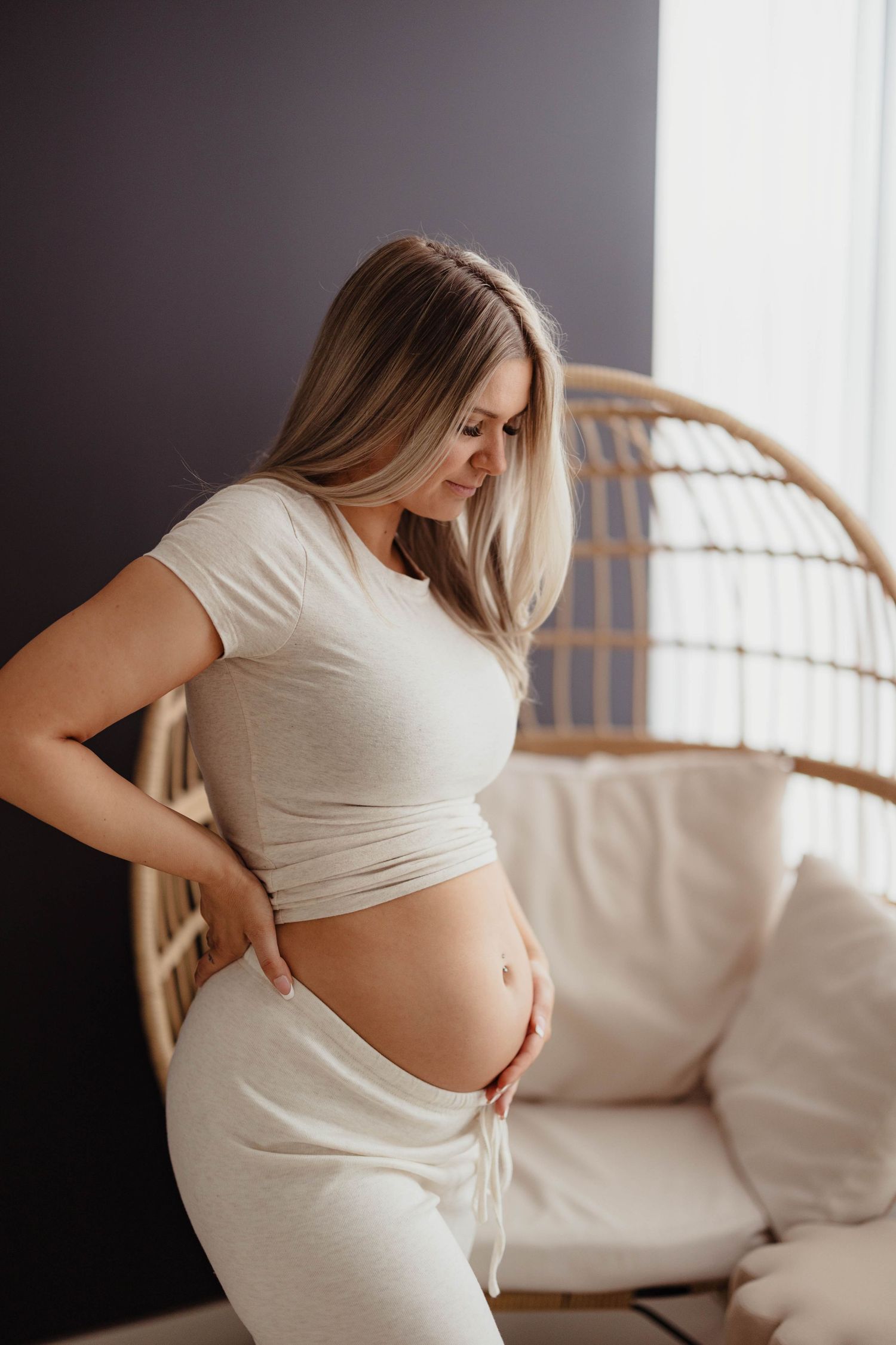 Pregnant woman in joggers holding baby bump beside a rattan egg chair near window. maternity pregnancy announcement maternity