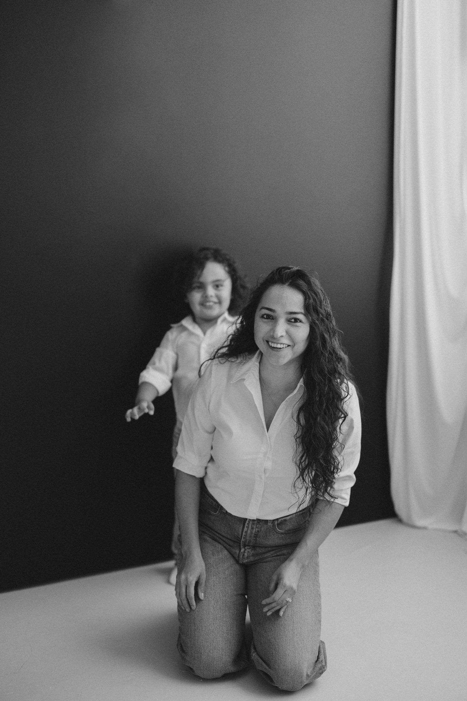 Black and white photo of smiling mother kneeling while young daughter poses behind her in studio. Lafayette indiana tippecanoe county