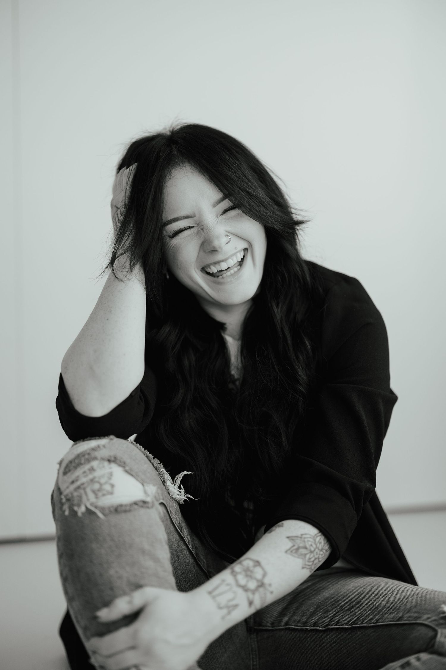 Black and white photo of laughing dark-haired woman with tattoos sitting on floor. Branding photos in west Lafayette Indiana