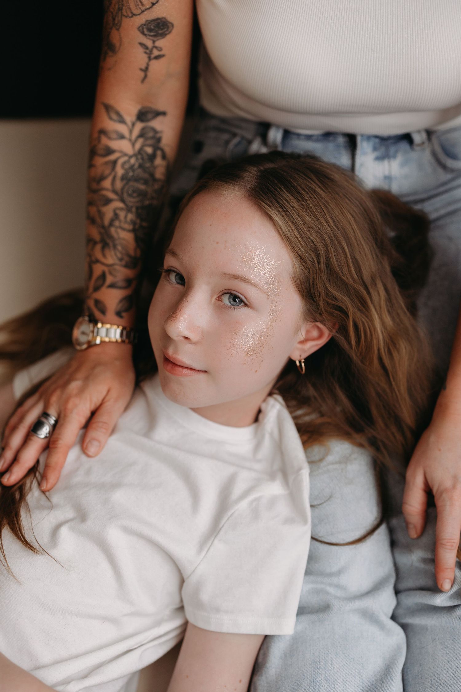 Young girl with freckles in white t-shirt leans against tattooed adult wearing jeans and a watch.