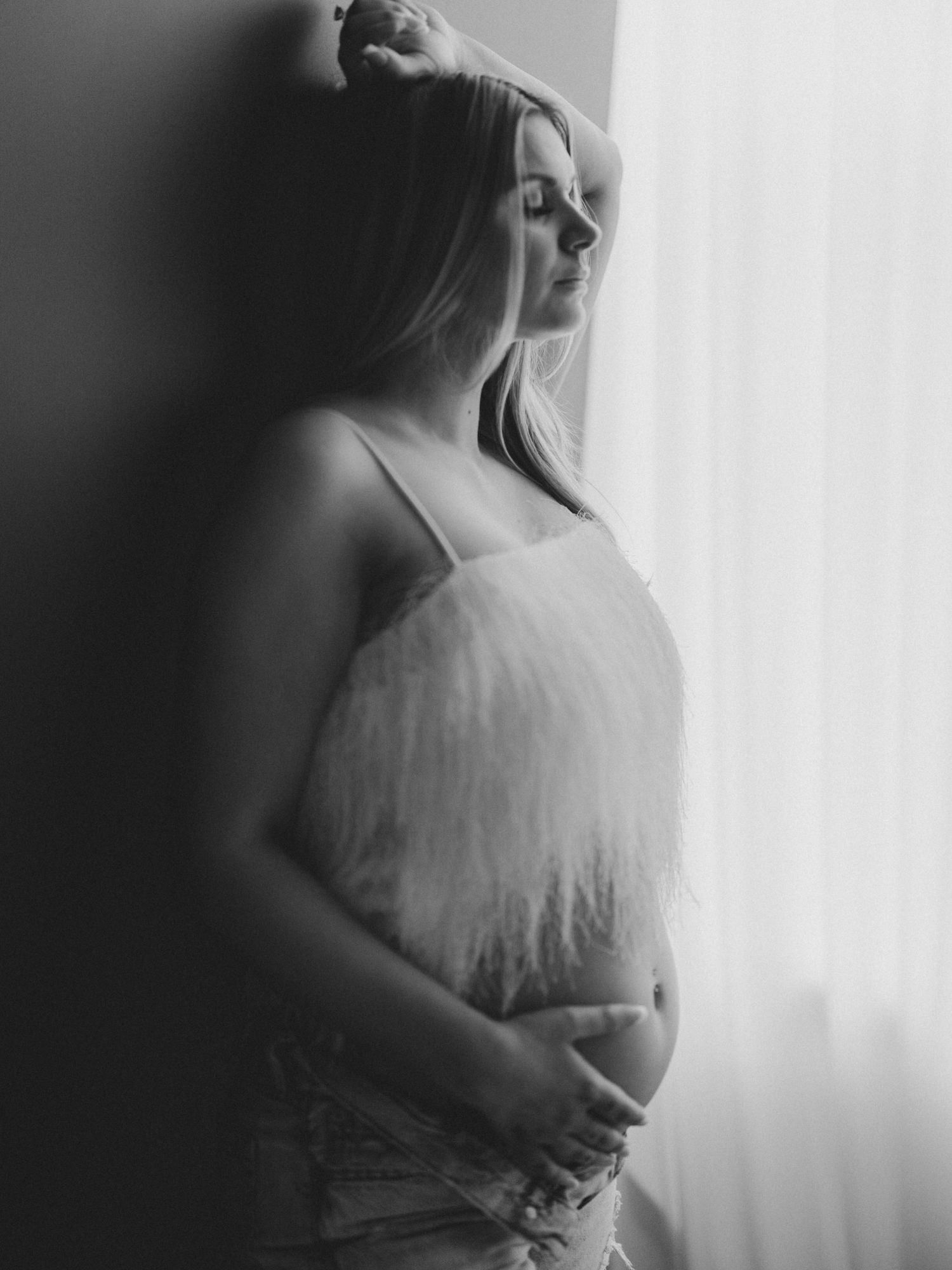 Black and white photo of pregnant woman in lace top leaning against wall holding baby bump by window. maternity lafayette