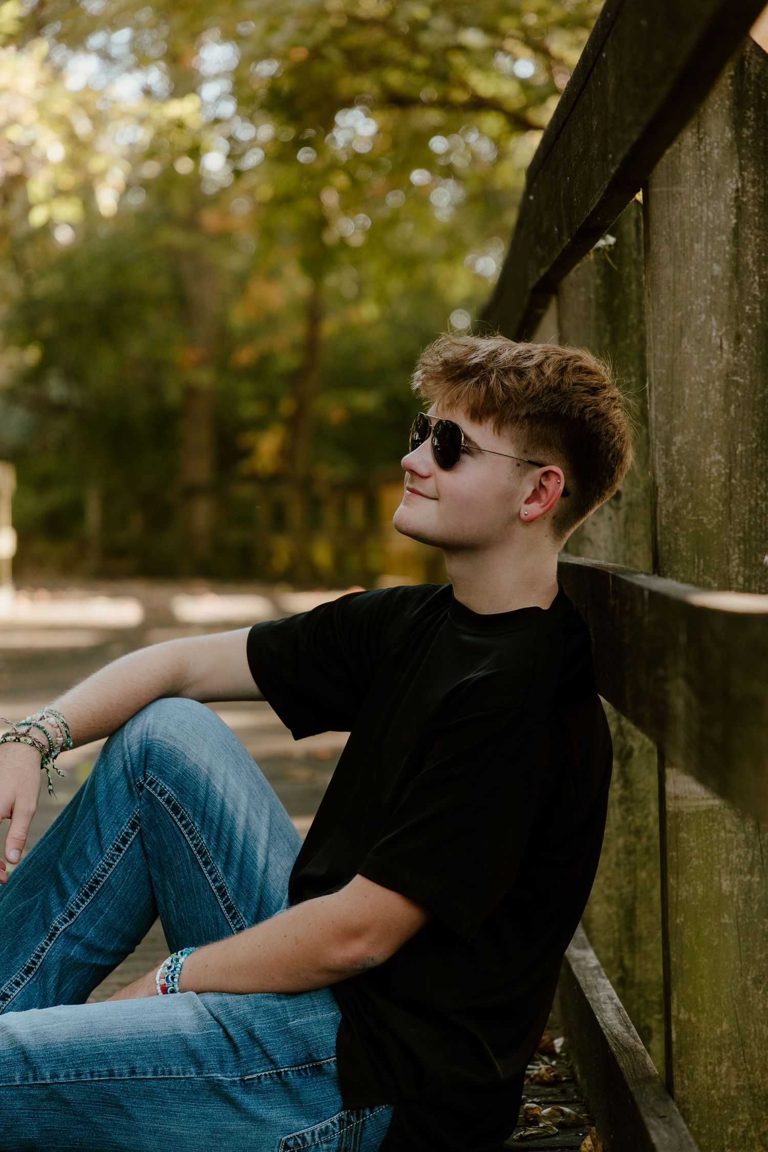 Teen boy in black shirt and sunglasses leaning against a wooden fence in autumn park. High school senior photos in west Lafayette indiana