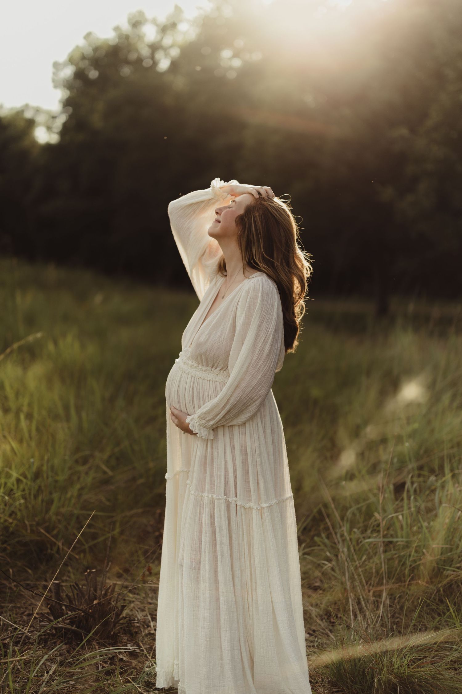 Pregnant woman in white boho dress standing in sunlit field, cradling baby bump at golden hour.