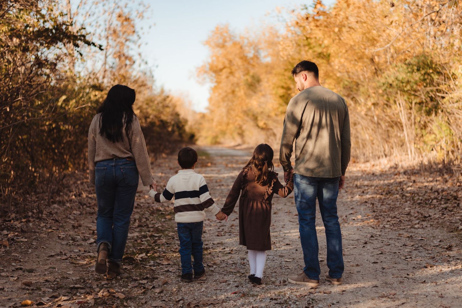 Family of four walking hand in hand down a sunlit autumn dirt path. Lafayette indiana tippecanoe county west lafayette
