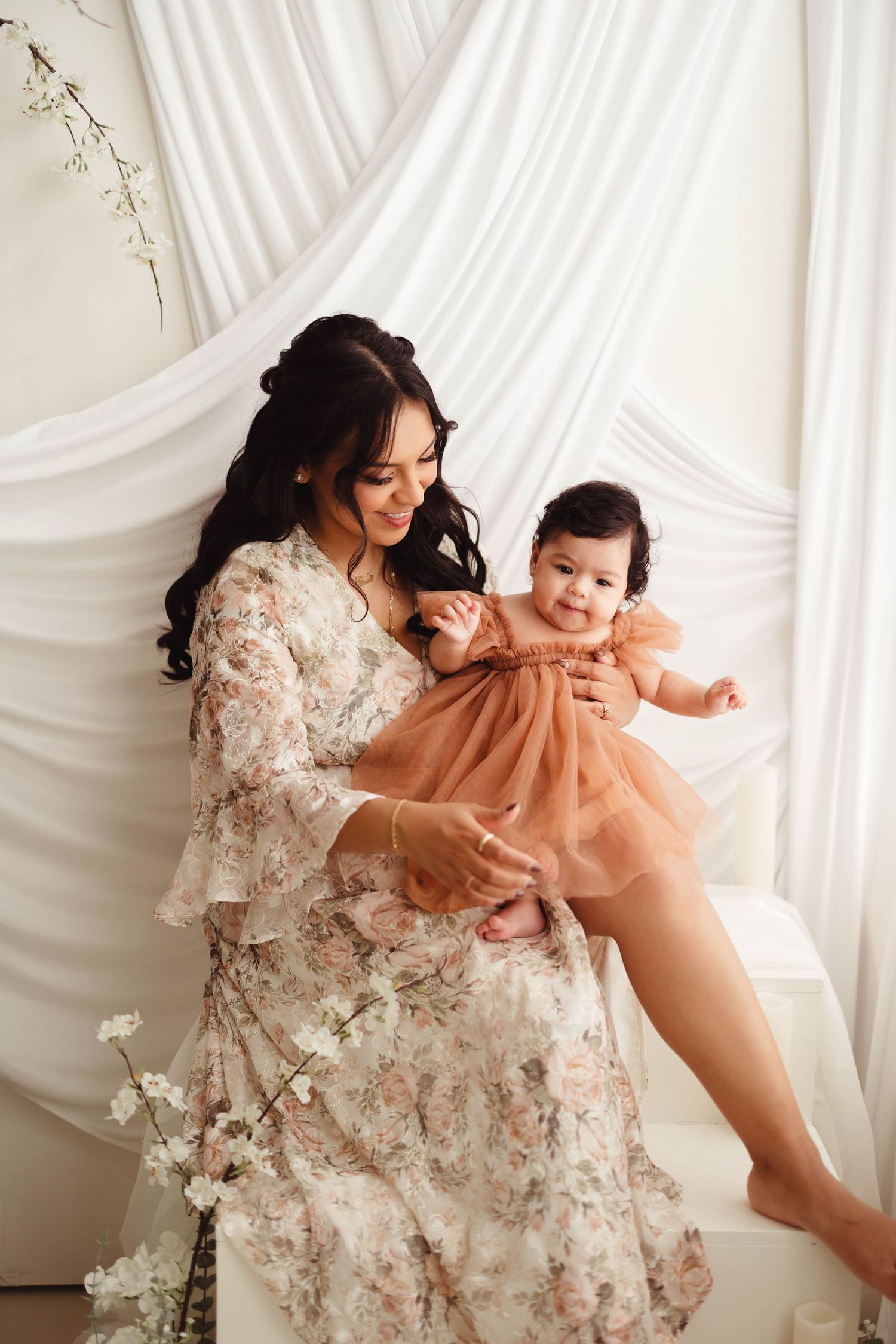 Mother in floral dress holds baby girl in peach tulle dress against white draped backdrop. Lafayette indiana tippecanoe county