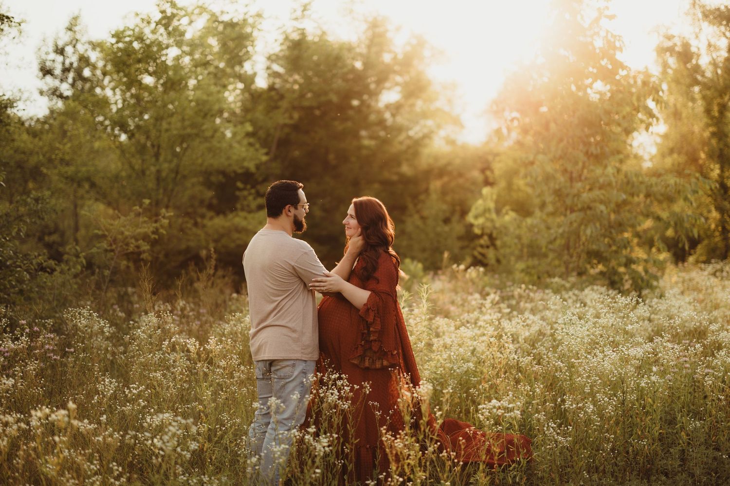 Couple holding hands in a sunlit wildflower meadow at golden hour, woman in rust dress, romantic maternity photo.