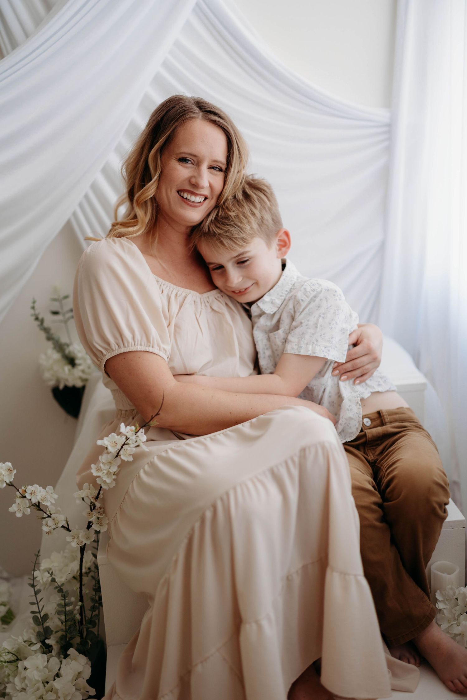 A smiling blonde mother in a cream dress sits with her young son hugging her warmly. in tippecanoe county indiana