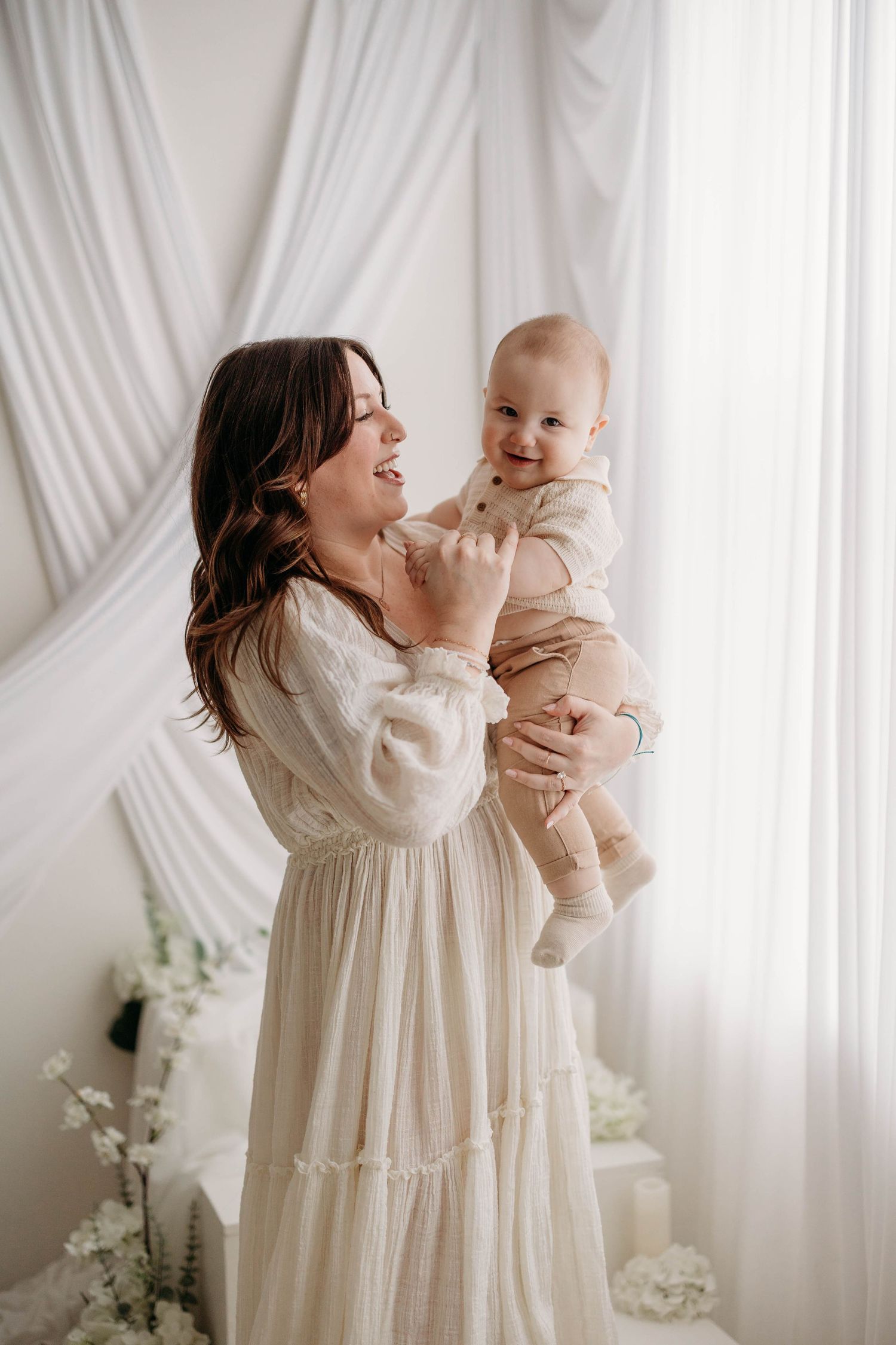 A smiling mother in a white dress holds her happy baby boy in a bright floral studio setting. mothers day photoshoot in Lafayette indiana