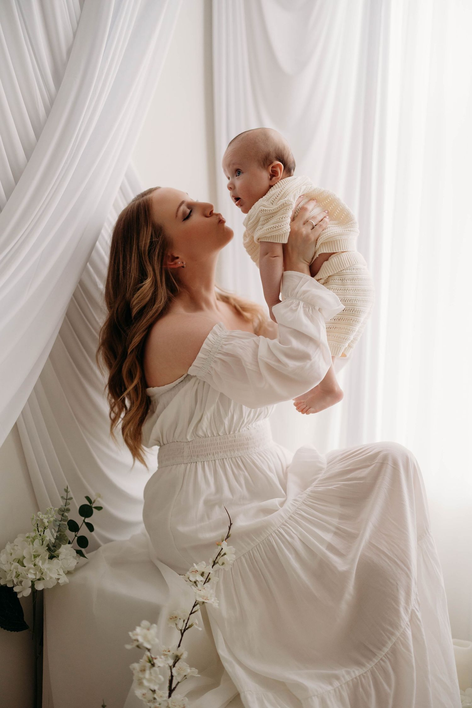 Elegant mother in white dress lifts newborn baby while seated near white curtains and flowers. Lafayette indiana tippecanoe county