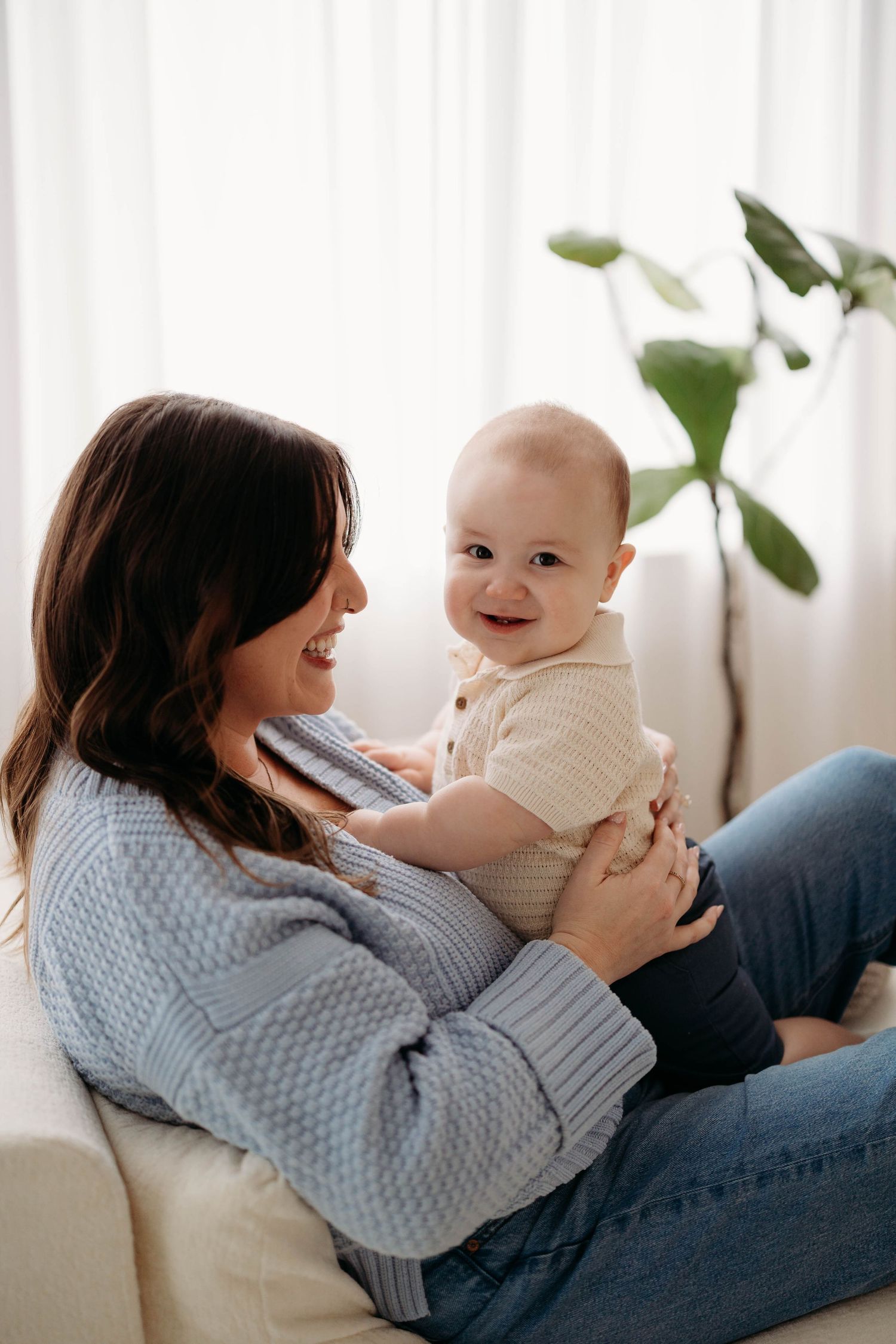 A smiling mother holds her happy baby on a couch near a bright window with a green plant. Lafayette indiana tippecanoe county west lafayette