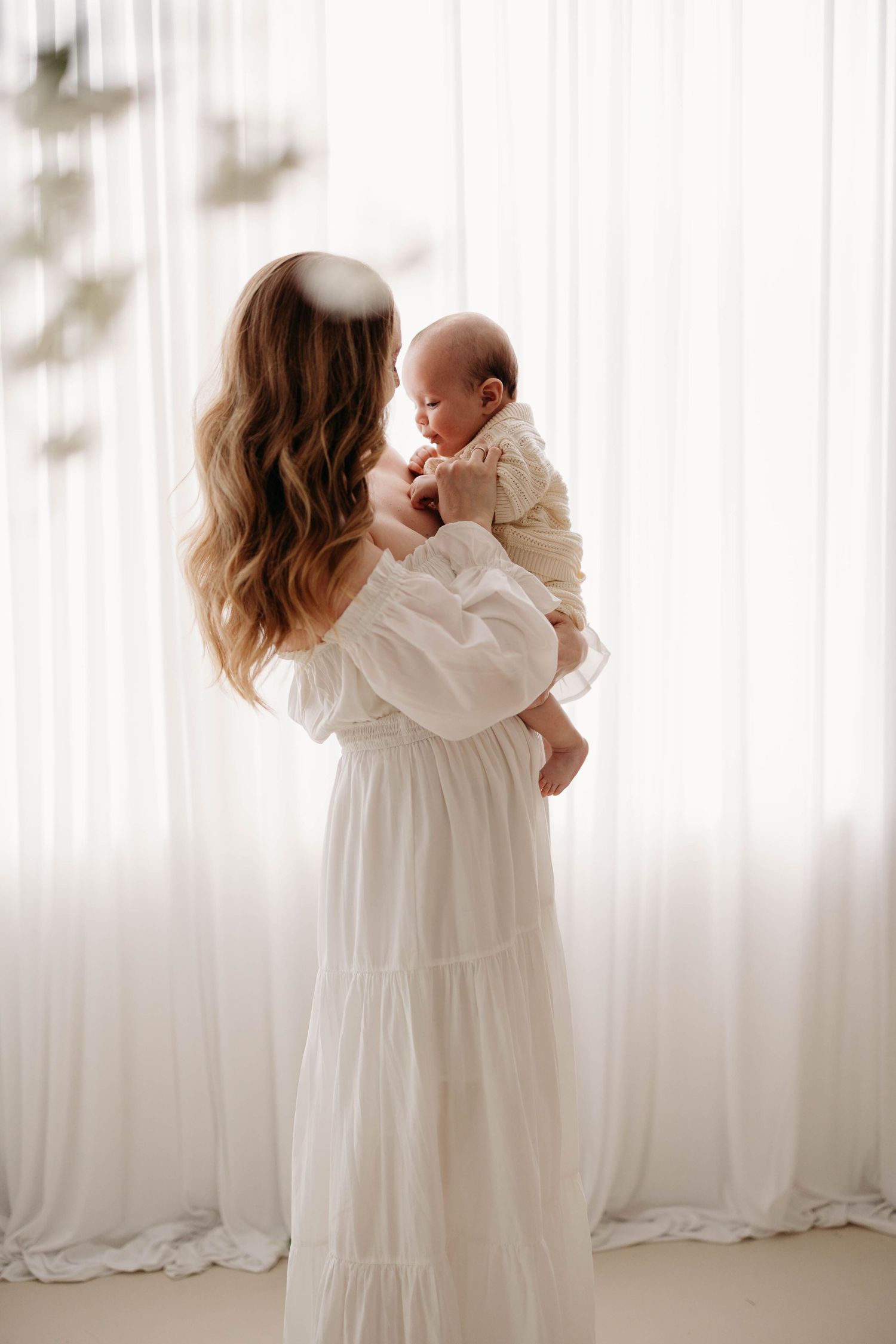 Mother in white dress holds newborn baby while standing before sheer white curtained window. Lafayette indiana west Lafayette
