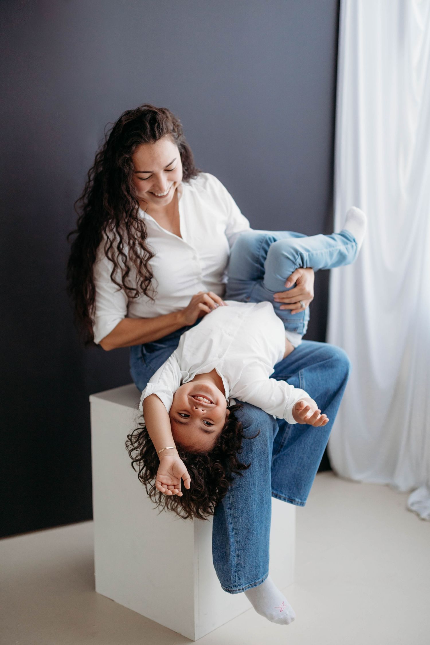 A playful mother with curly hair laughs as her child hangs upside down from a white cube. Lafayette indiana tippecanoe county west lafayette