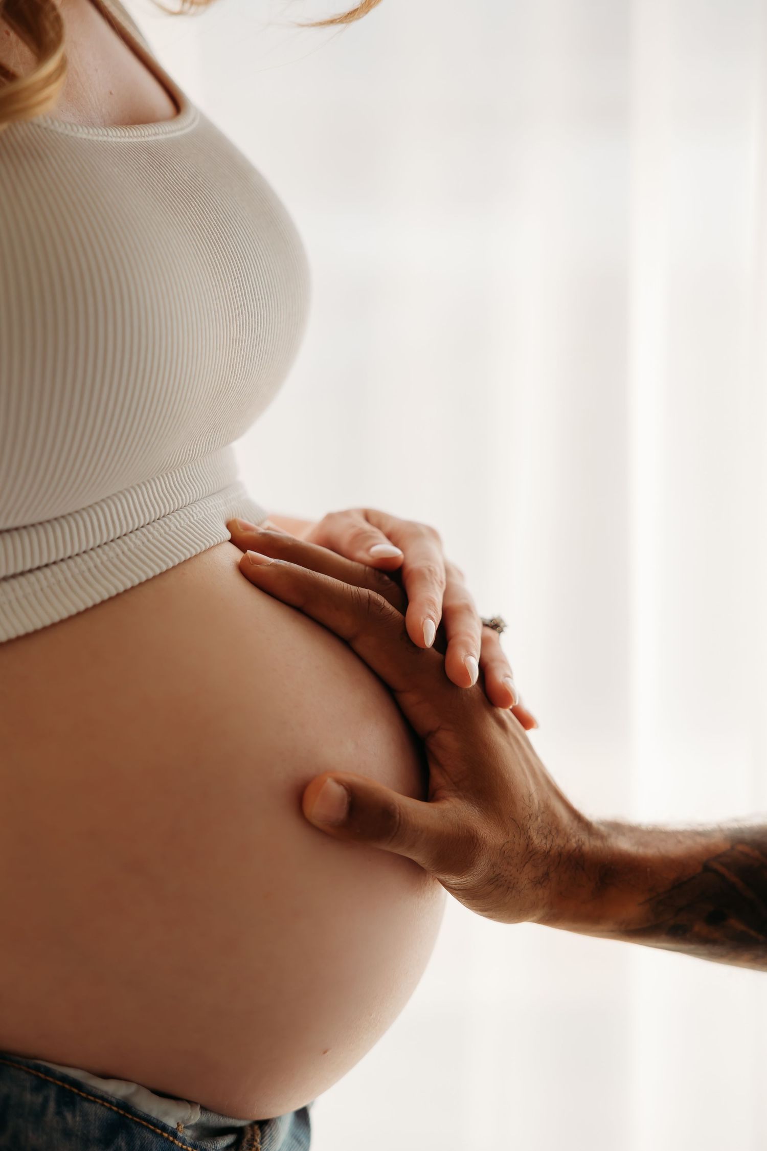 A pregnant woman in a white crop top cradles her baby bump as a tattooed hand gently rests on it. Lafayette indiana tippecanoe county west l