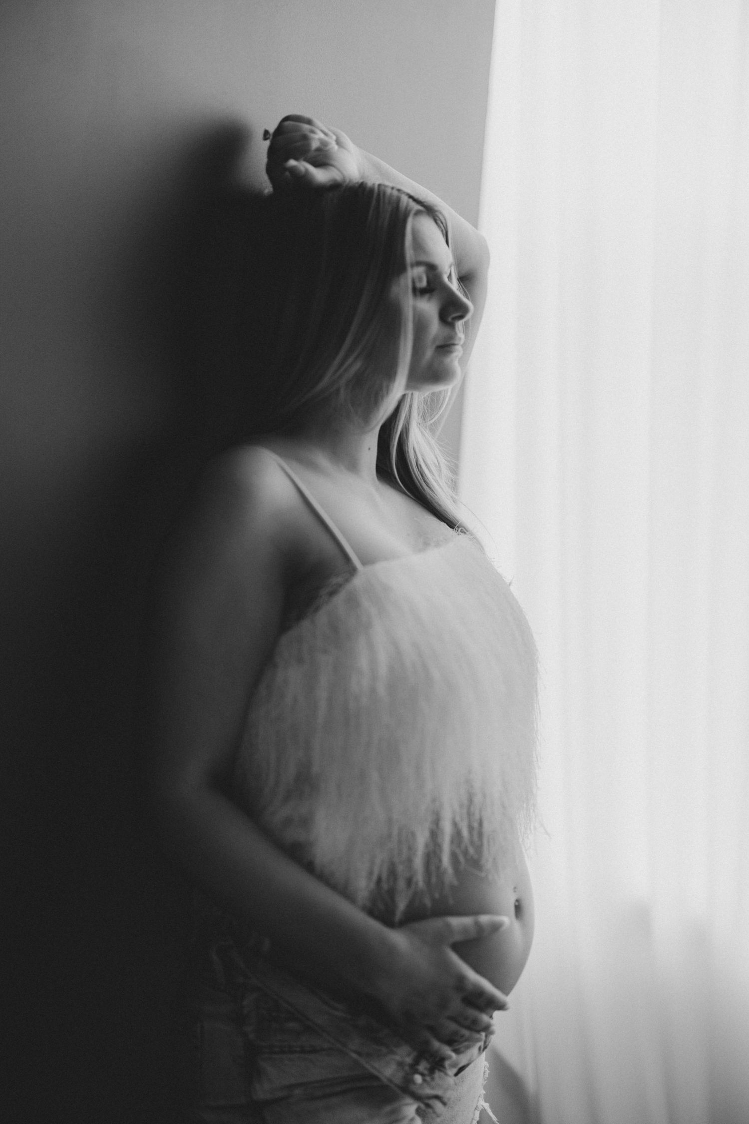 A black and white portrait of a pregnant woman holding her belly near a curtained window. Lafayette indiana tippecanoe county west lafayette