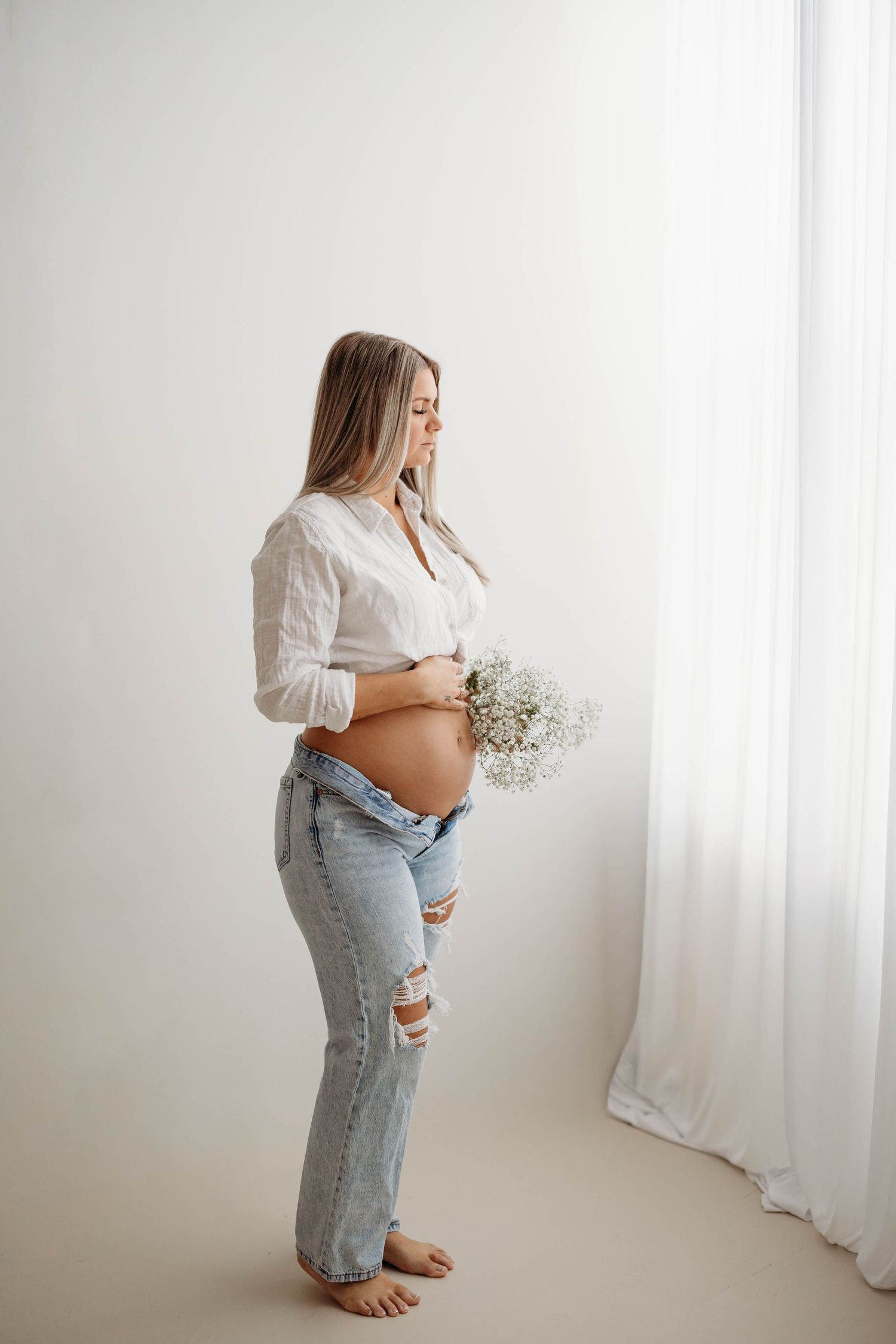 Pregnant woman in white shirt and ripped jeans holding baby's breath flowers against white curtain backdrop. maternity Lafayette indiana