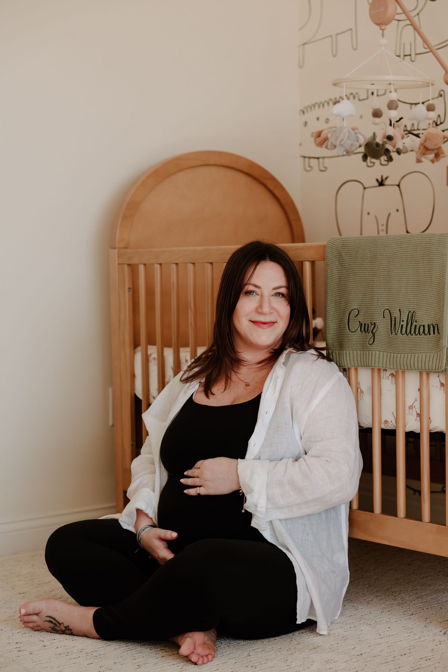 Pregnant woman in black outfit sitting cross-legged on floor beside wooden crib in nursery. in home maternity session neutral
