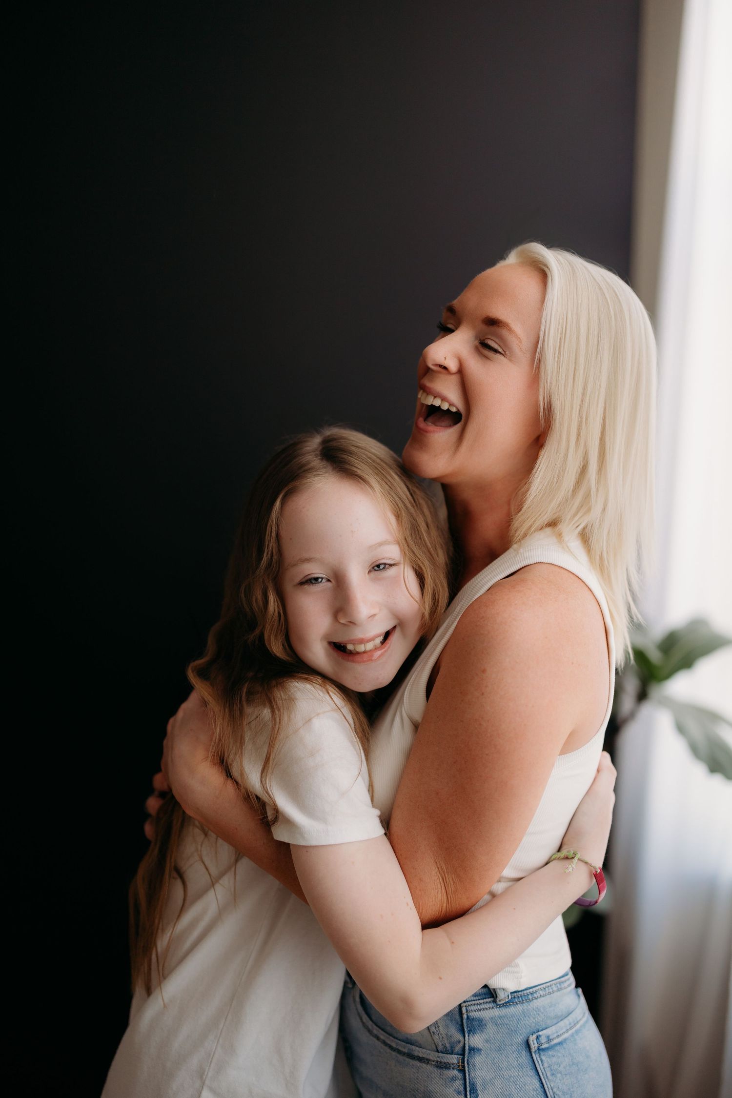 A laughing blonde woman hugs a smiling young girl in white shirts against a dark wall.