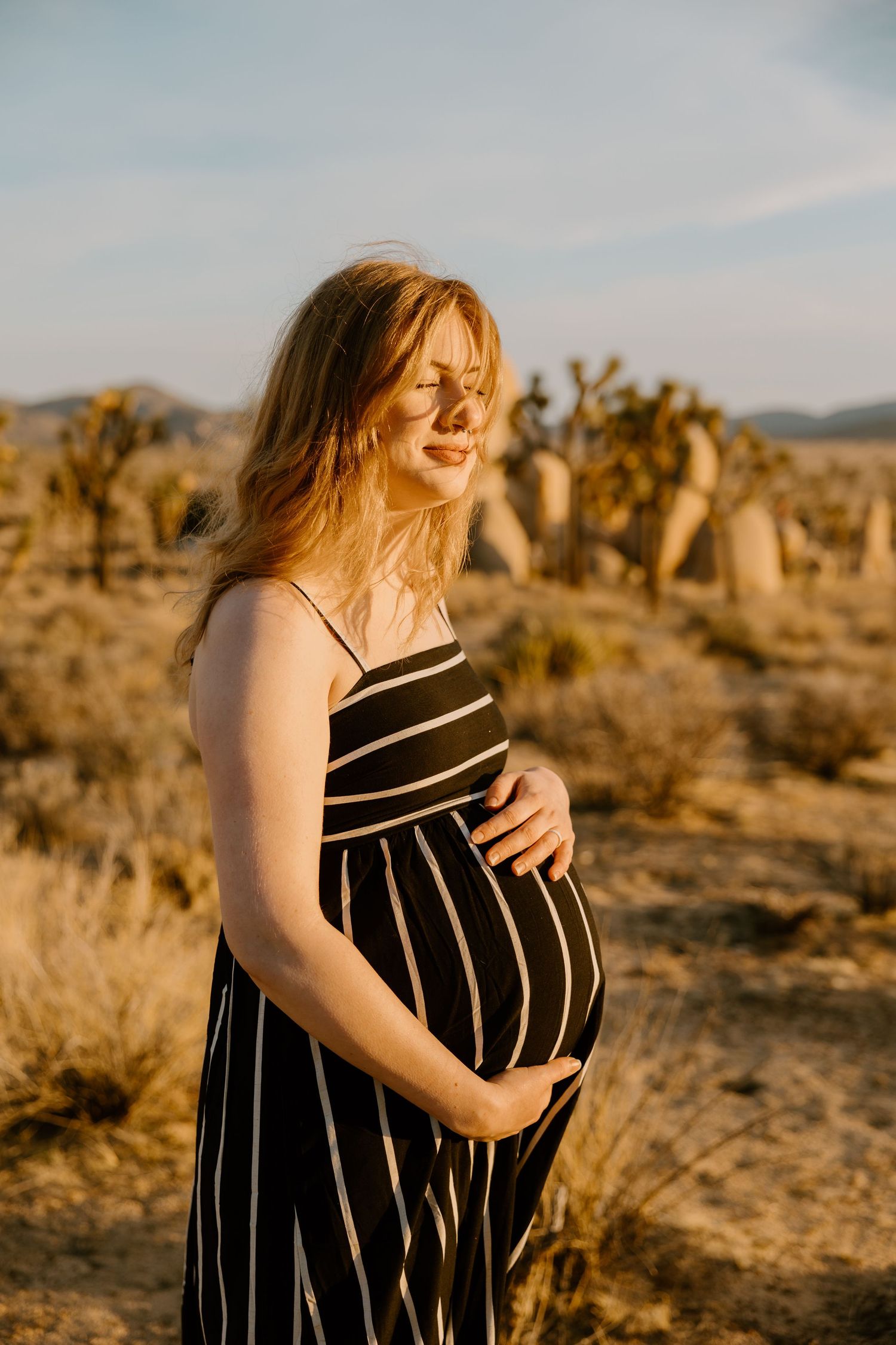 Pregnant woman in striped maxi dress cradling baby bump in golden desert landscape at sunset. Lafayette indiana tippecanoe county