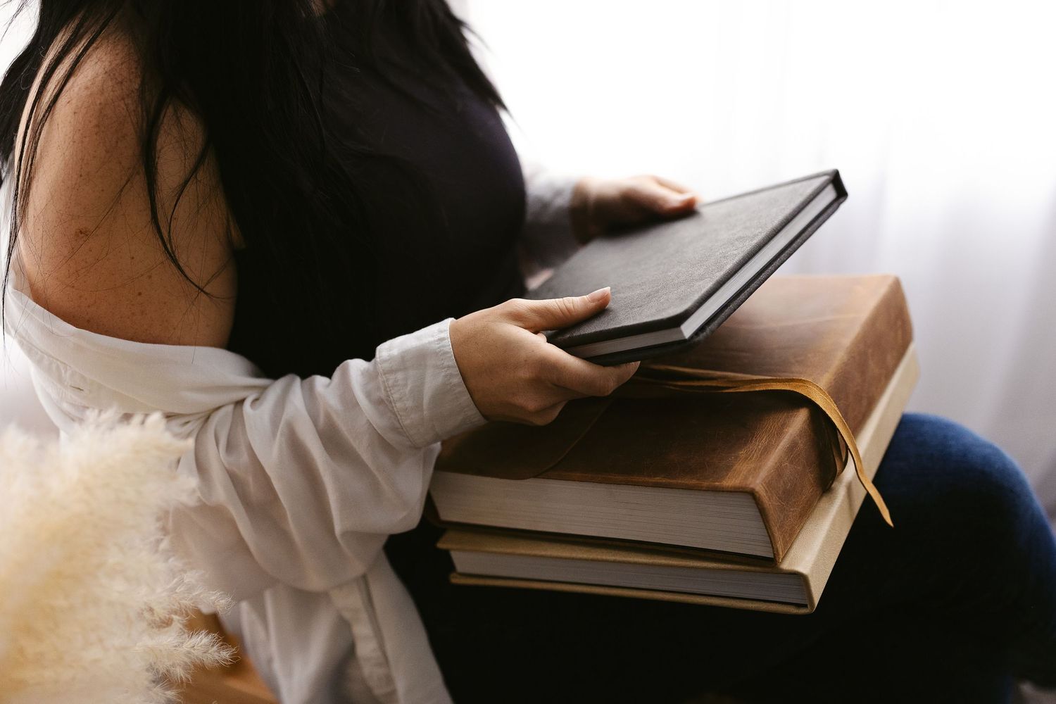 Woman with dark hair holding leather-bound photo albums and books while seated by window.