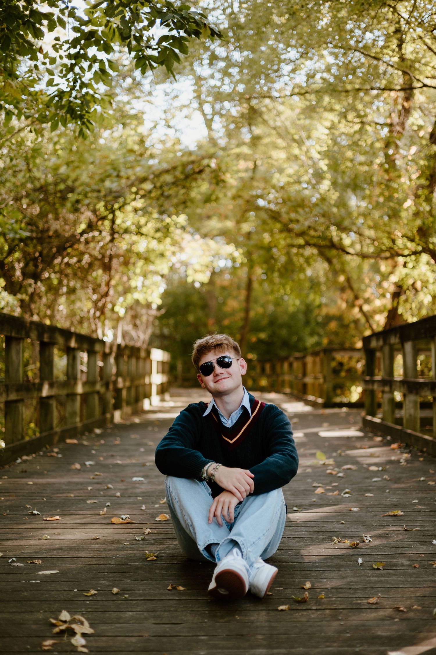 Young man in sunglasses and dark sweater sitting relaxed on a wooden bridge in autumn. High school senior photos in west Lafayette indiana