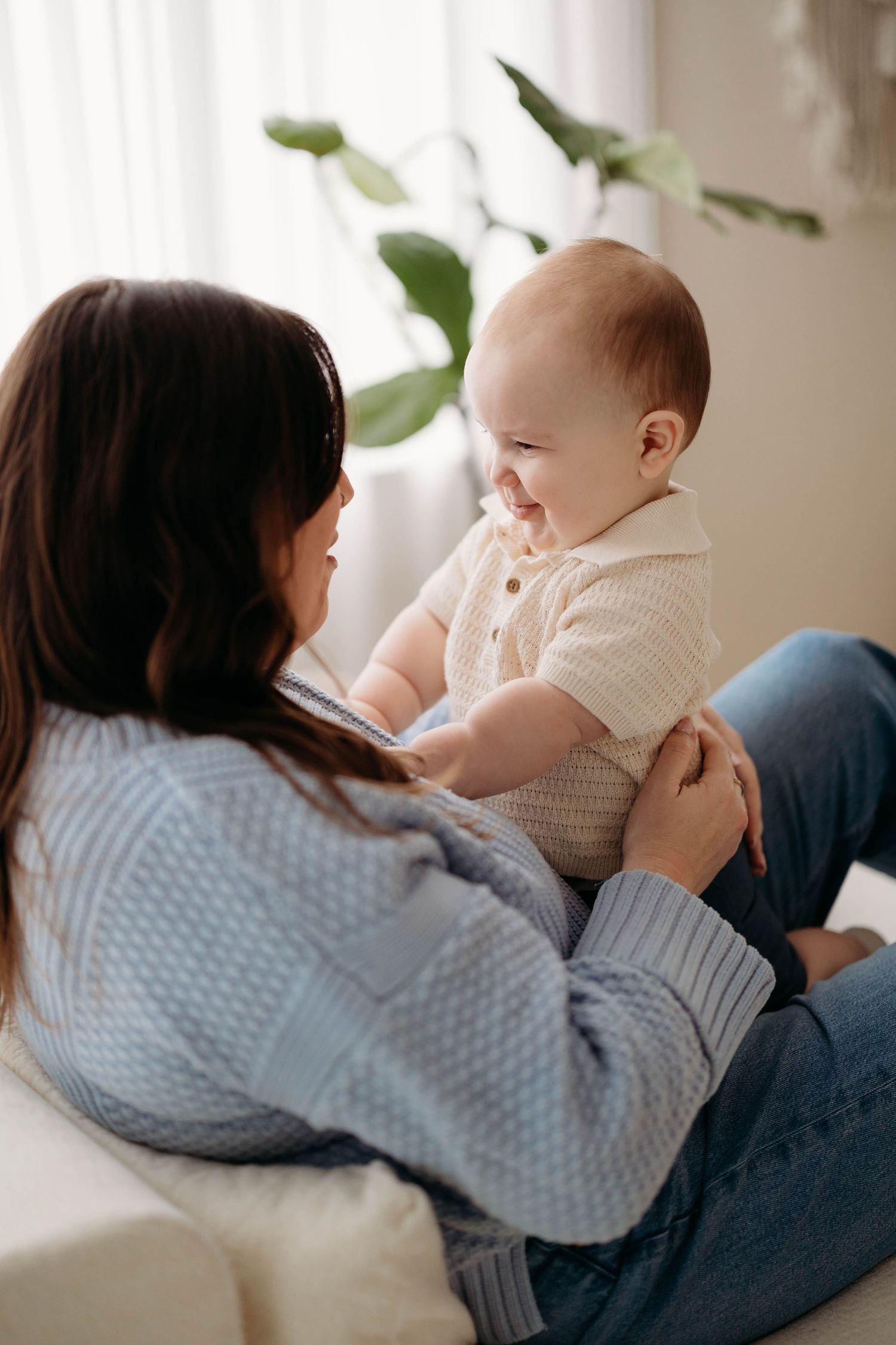 A loving mother in a blue sweater holds her smiling baby while sitting on a white couch. Lafayette indiana tippecanoe county west lafayette