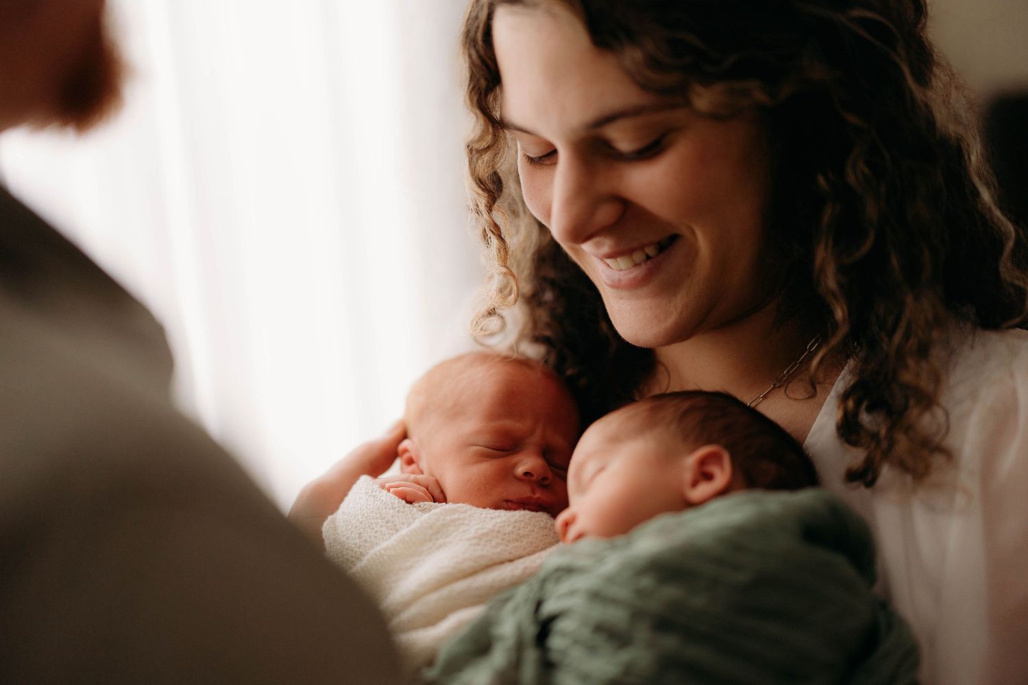 Smiling mother with curly hair tenderly holding two sleeping newborn twins near a window. Lafayette indiana tippecanoe county west lafayette