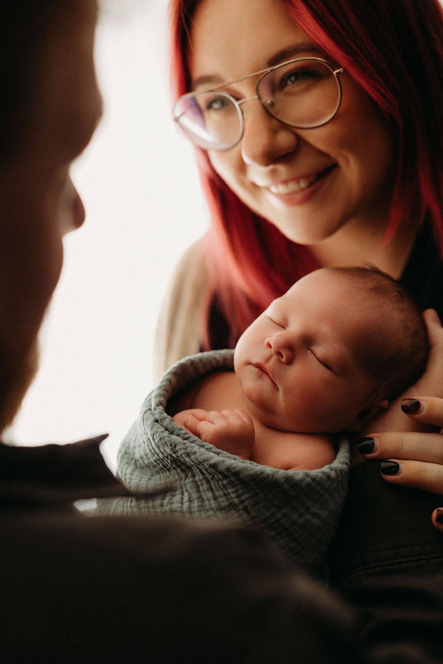 Smiling red-haired woman with glasses holding a sleeping newborn baby wrapped in a blue blanket.