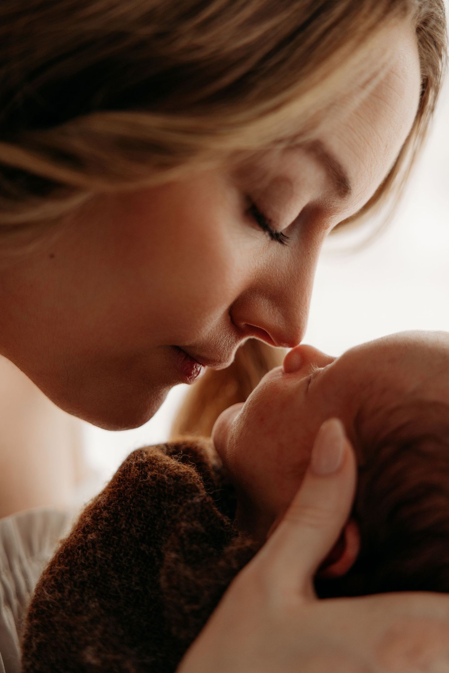 A tender mother gently kisses her newborn baby wrapped in a brown blanket in a warm, softly lit portrait.