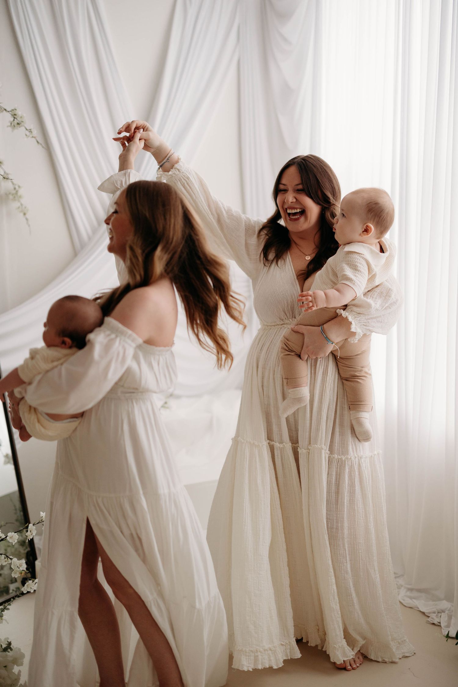 Two joyful mothers in white dresses dancing and holding their babies in a bright studio. Lafayette indiana tippecanoe county west lafayette