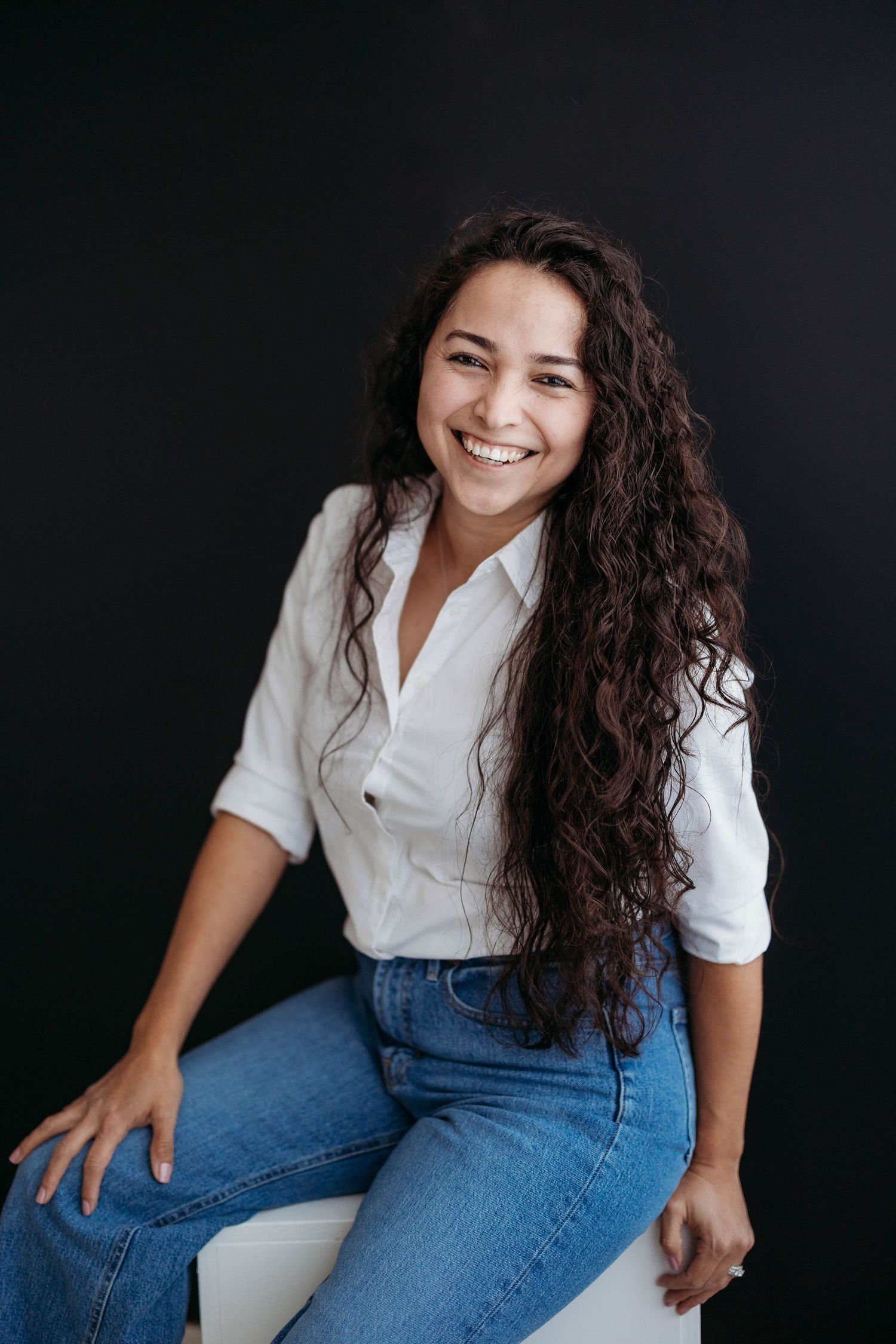 A smiling young woman with long curly dark hair sits confidently in a white shirt and jeans. Lafayette indiana tippecanoe county