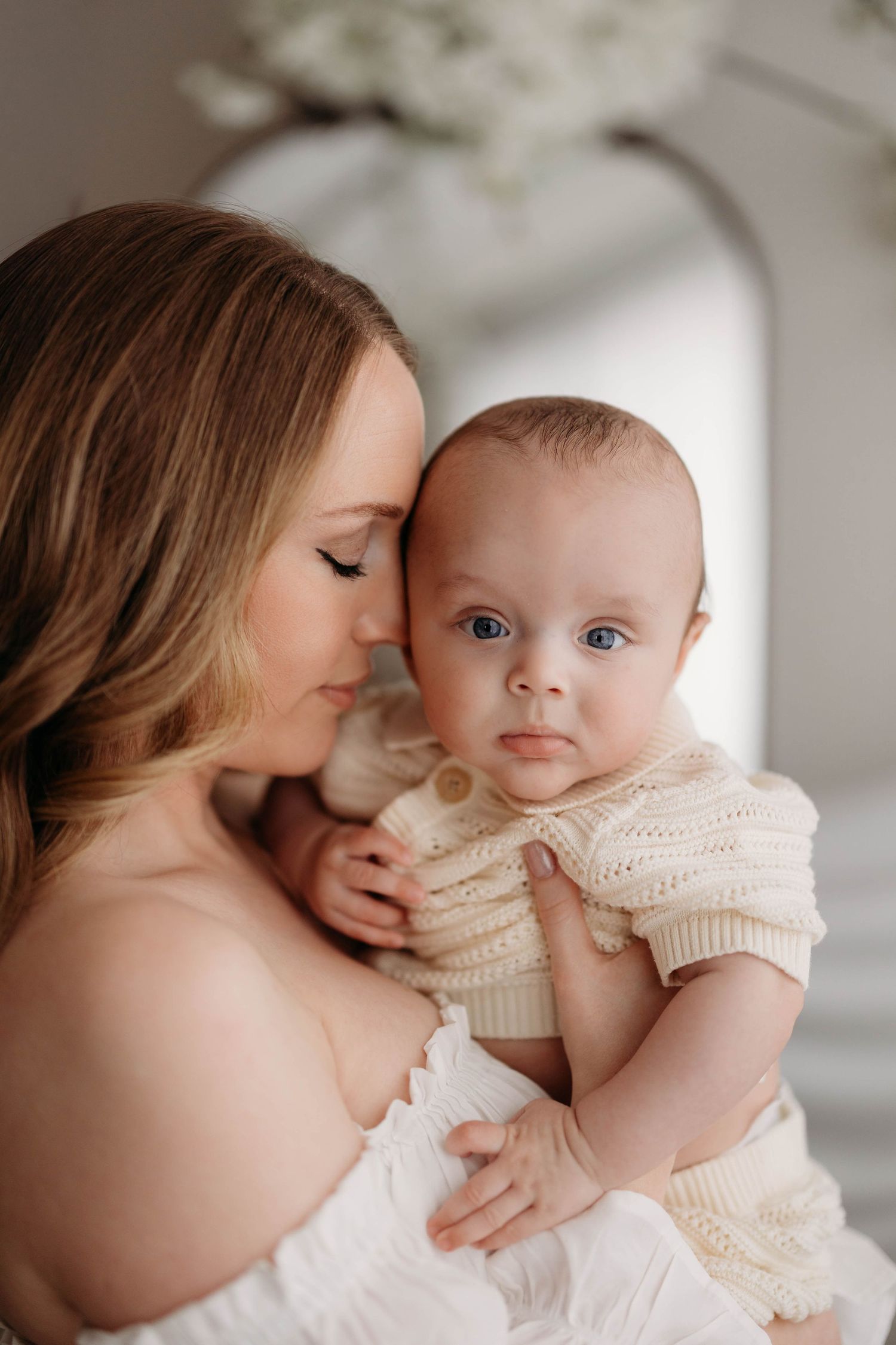 Mother in white off-shoulder dress cuddles blue-eyed baby in cream knit outfit indoors. Lafayette indiana tippecanoe county west lafayette