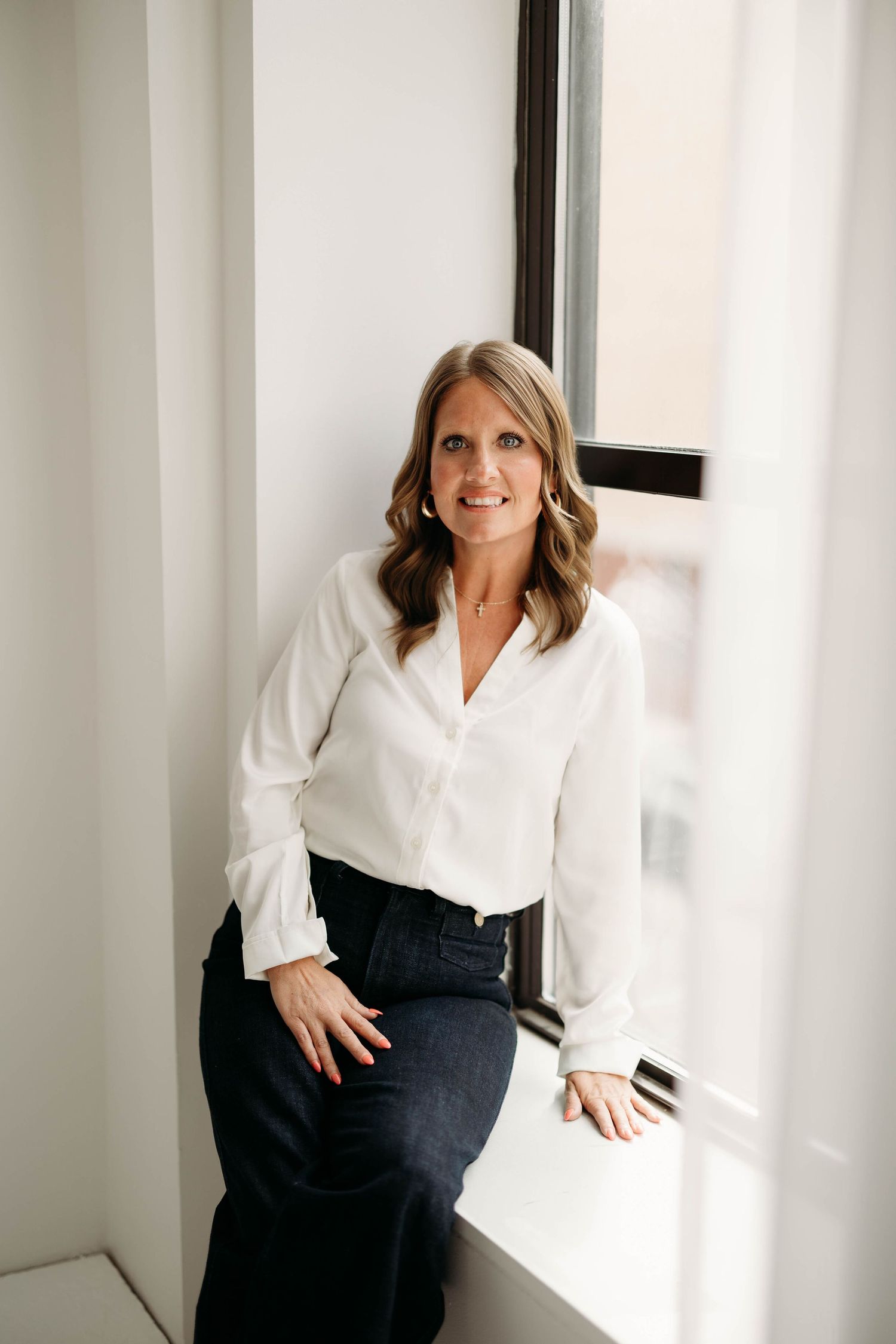 Professional woman in white blouse and dark pants leaning against window in bright office. Branding photos in west Lafayette Indiana