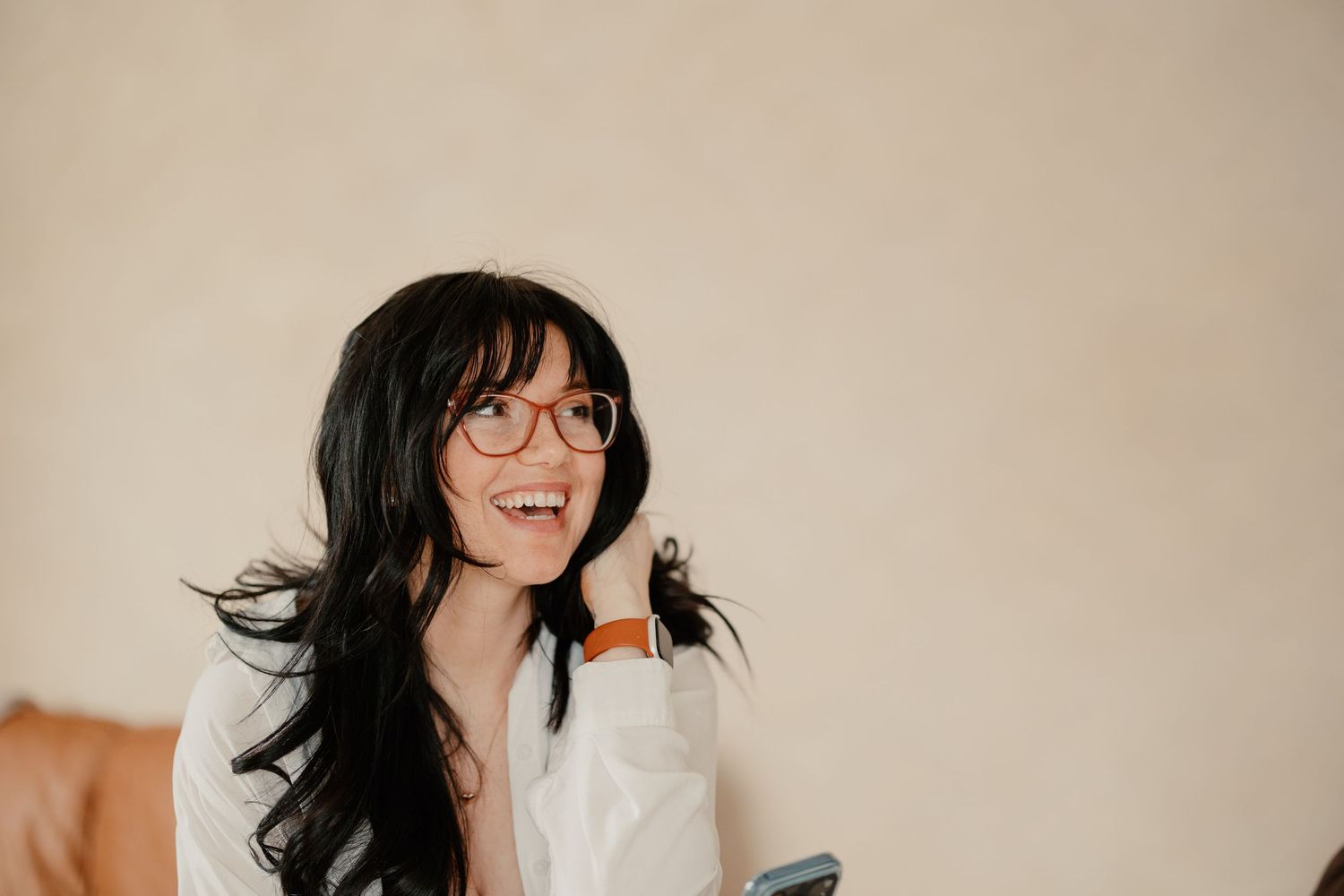 Smiling young woman with long dark hair and round glasses wearing a white shirt, looking up joyfully indoors.